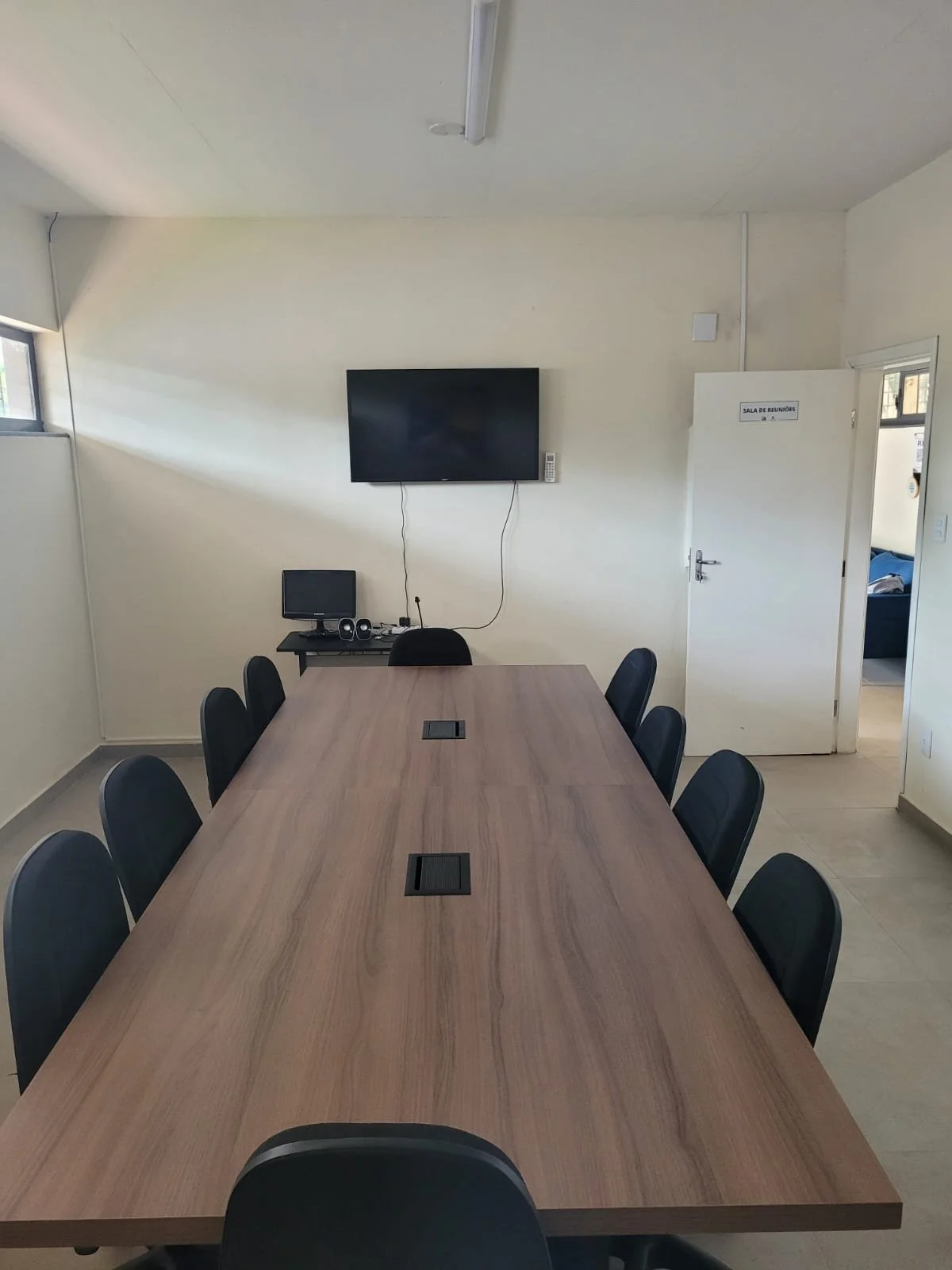 Sala de reuniões com mesa retangular, cadeiras, televisão e equipamentos de videoconferência.