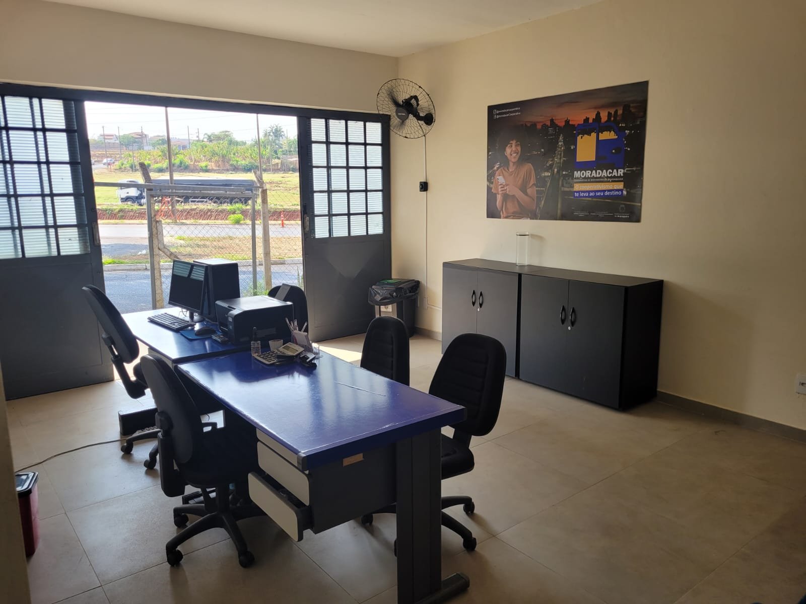 Escritório com mesa azul, computadores, cadeiras giratórias, armários e pôster da MoradaCar.