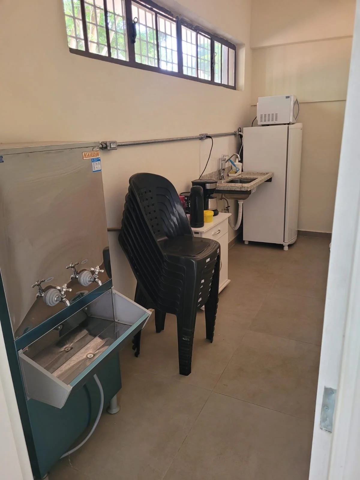 Área de serviço com geladeira, pia, micro-ondas, bebedouro industrial e cadeiras plásticas empilhadas.