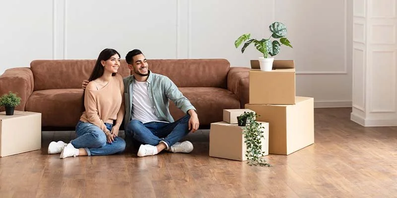 A smiling young couple sitting on the floor in a living room with moving boxes and potted plants.