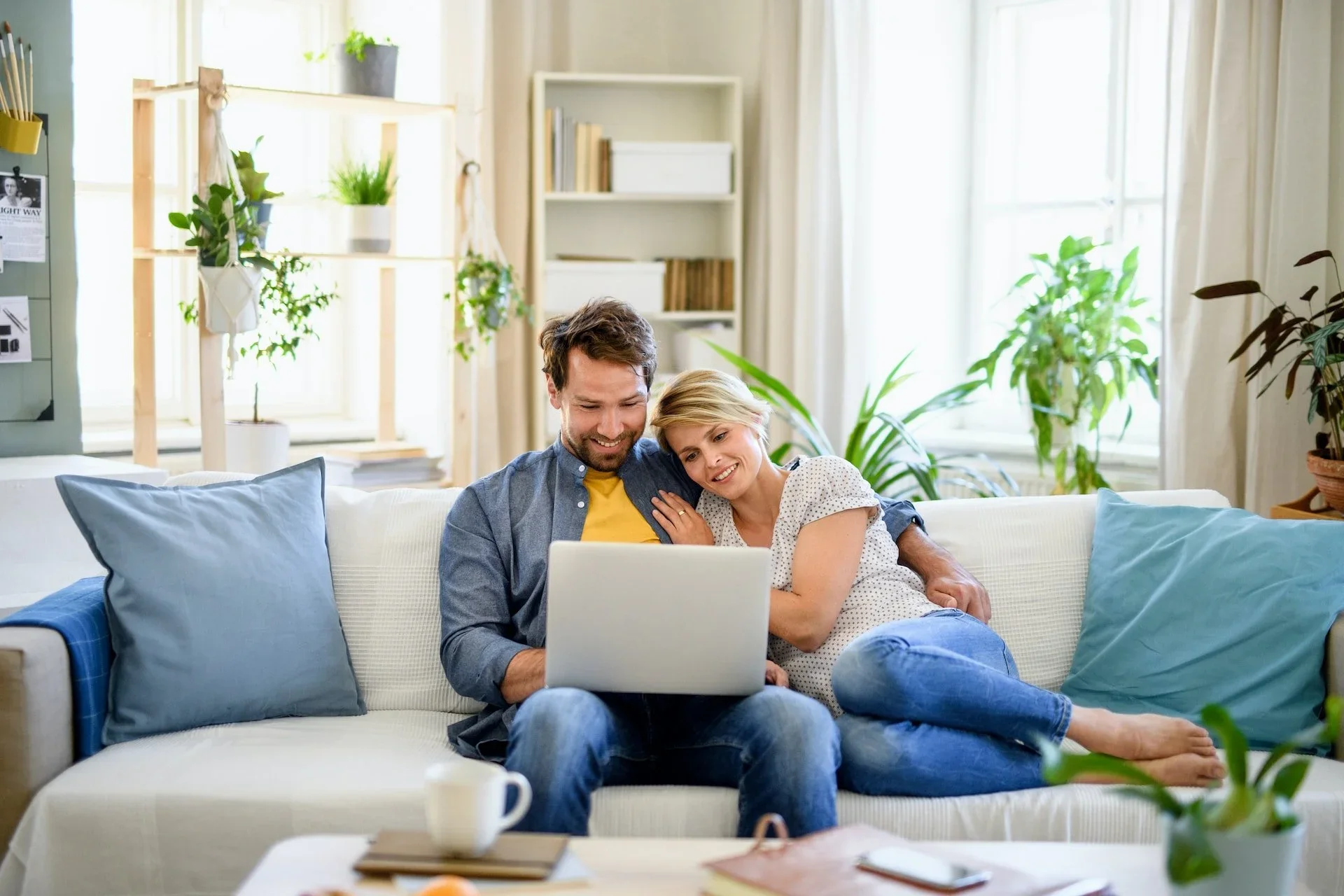 A happy couple sitting on a white sofa, looking at a laptop, surrounded by pillows, in a bright living room.