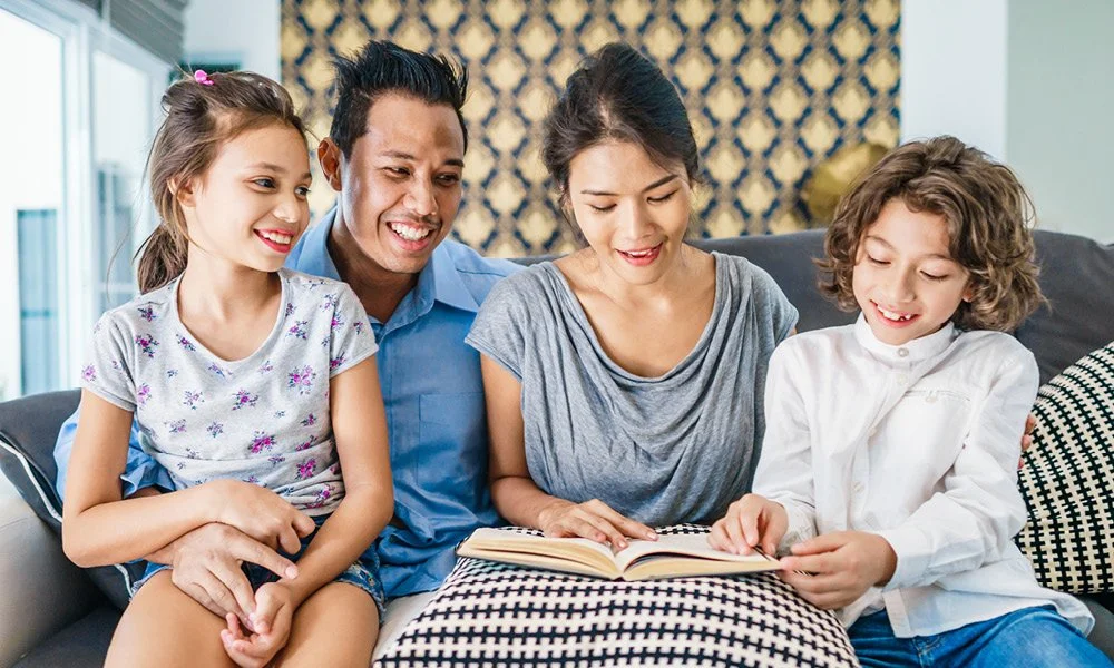 A family of four sitting on a couch, reading a book together and smiling.