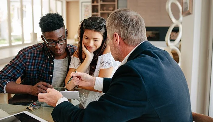 A man in a blazer showing something on his phone to a teenage boy and girl sitting at a table inside a bright room.