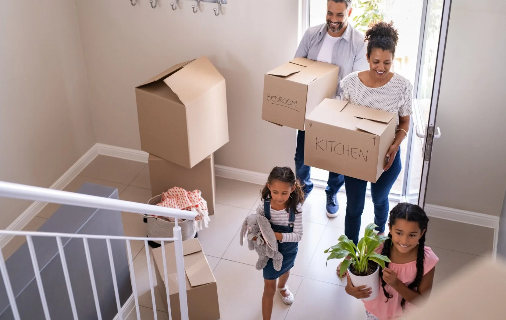Family moving into a new home, carrying cardboard boxes labeled bedroom and kitchen, with two young girls, one holding a potted plant and the other carrying a stuffed rabbit toy.