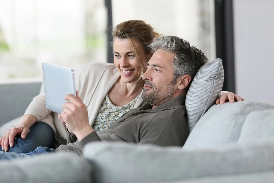 A woman and a man sitting on a couch, looking at a tablet together and smiling.