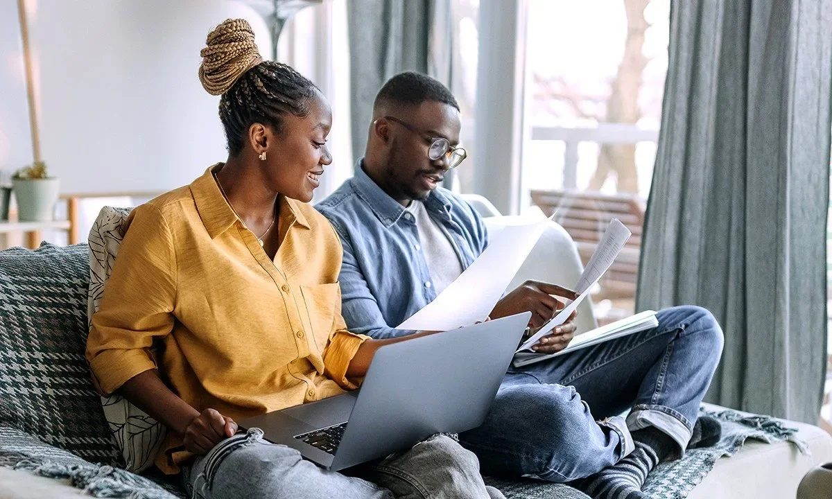 A man and woman sitting on a couch, looking at papers together with a laptop open in front of the woman, in a bright room with large windows and curtains.