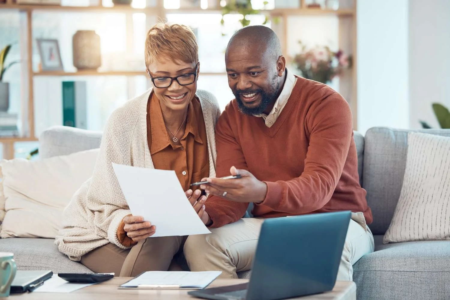 A middle-aged woman and man sitting together on a gray couch, reviewing documents and sharing a smartphone, with a laptop on the table in front of them in a bright living room.