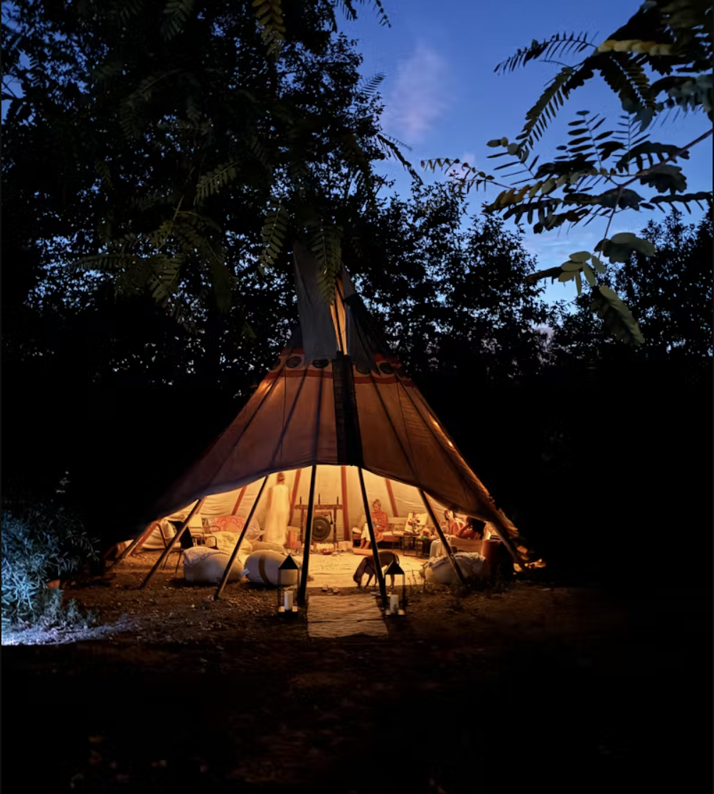 A warmly lit teepee tent set up outdoors at dusk, surrounded by trees and bushes, with people sitting inside and a pathway leading to the entrance.