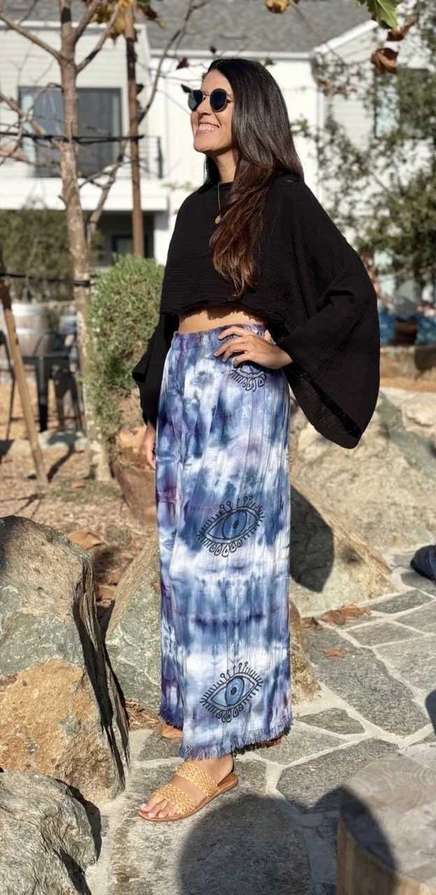 Woman wearing black sunglasses, black crop top, tie-dye skirt with eye design, and tan sandals standing outside near rocks and trees.
