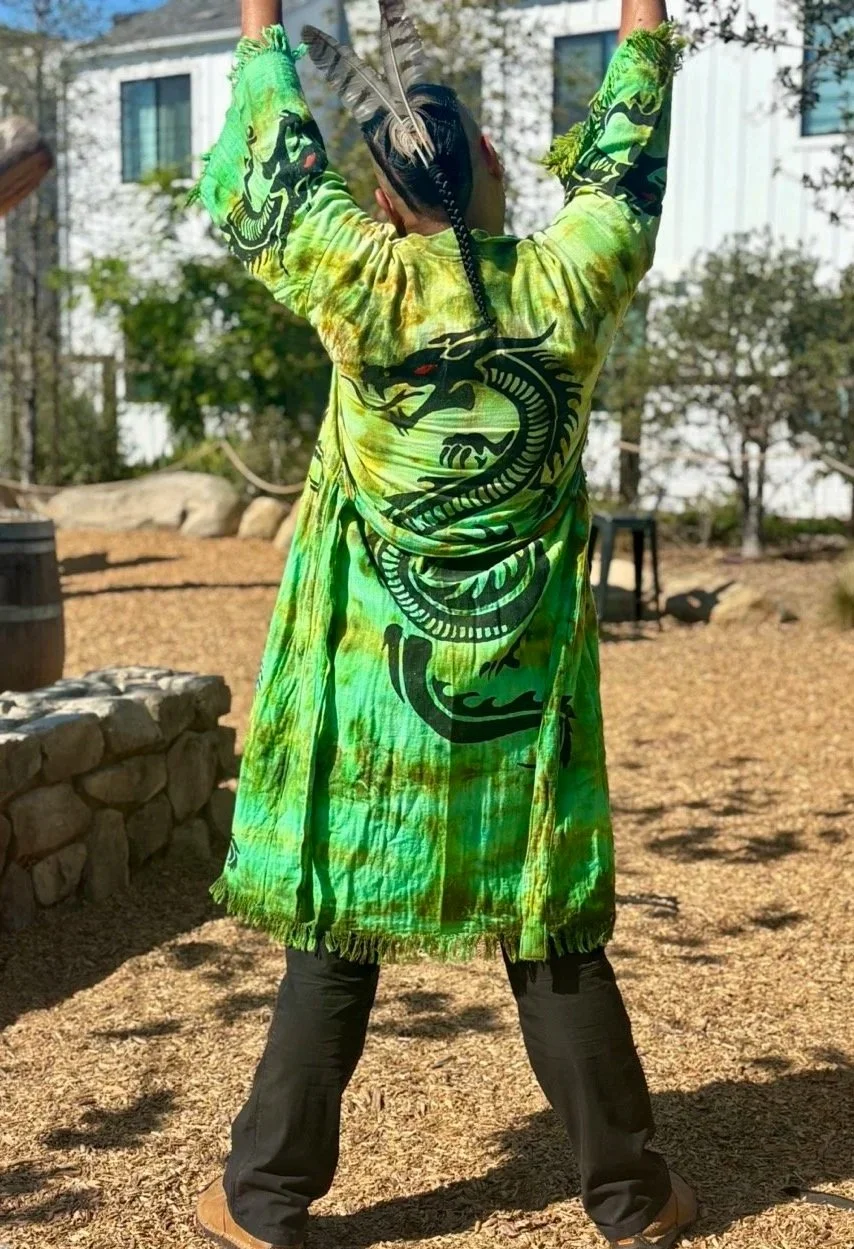 Person standing outdoors with arms up, wearing a vibrant green tie-dye coat with black dragon prints, black pants, and brown shoes, with a house and trees in the background.