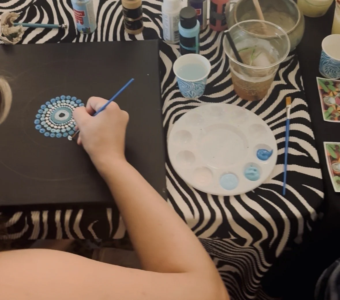 Person creating a dot mandala artwork on black paper with blue and white paint, surrounded by paint bottles, brushes, drinks, and art supplies on a zebra print tablecloth.