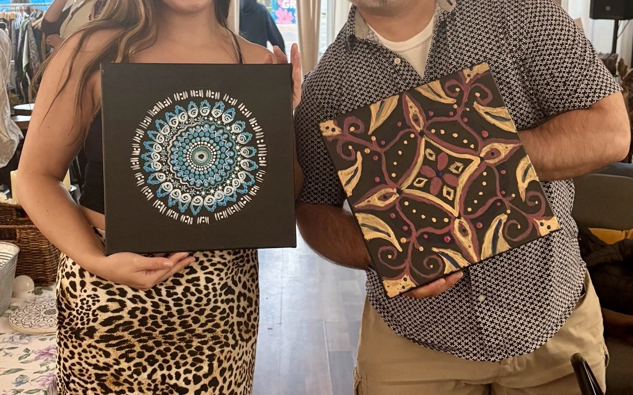 Two people holding up colorful patterned canvases, one with a mandala design in blue, white, and black, and the other with an abstract floral design in red, yellow, and black.