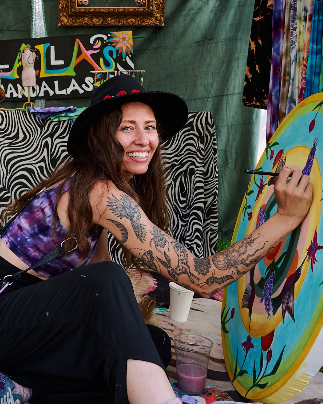 A woman with tattoos and a black hat painting a colorful, floral-themed artwork on a canvas.