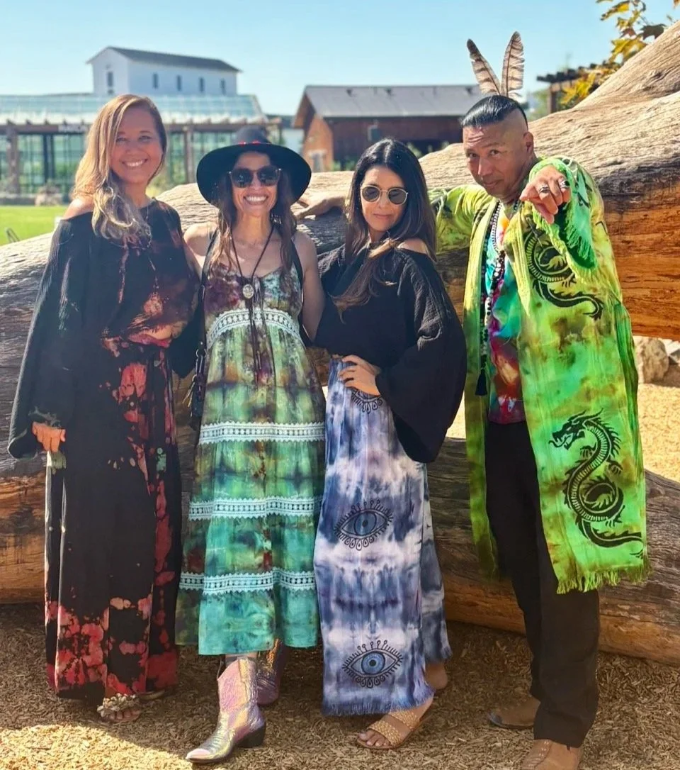 Four people outdoors on a sunny day, two women in long colorful dresses and a man in a bright green jacket with dragon designs, all smiling and posing in front of a large fallen tree.