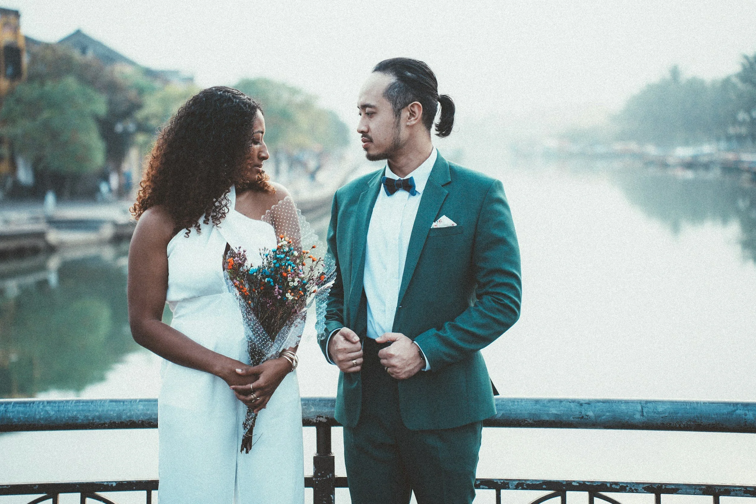 An interracial couple dressed in formal attire standing closely by a river with a blurred background of trees and buildings.