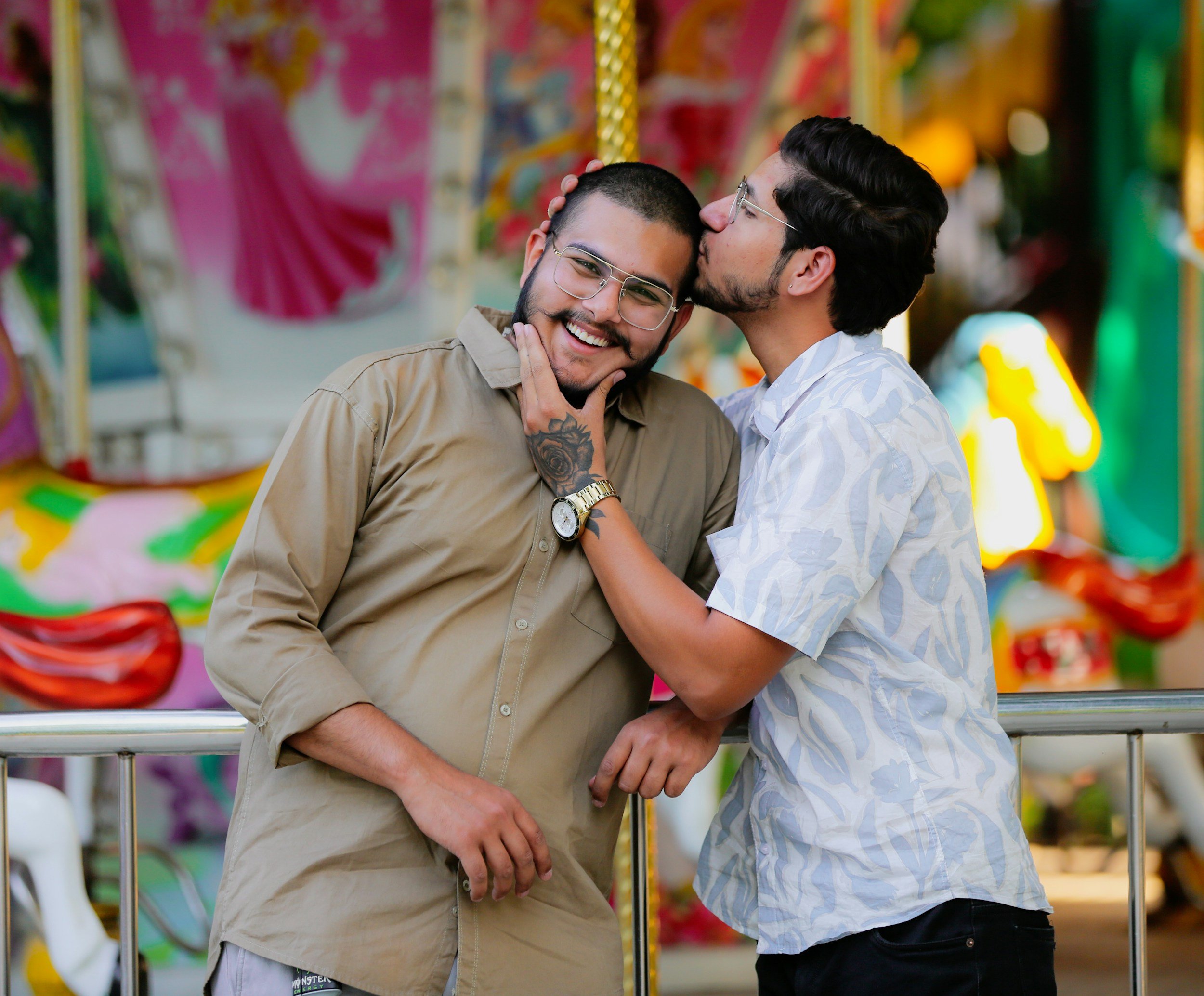 Two young men sharing a moment of affection at an amusement park or fair, one kissing the other's head while the other smiles warmly, both wearing glasses and casual clothing, with colorful rides and attractions blurred in the background.