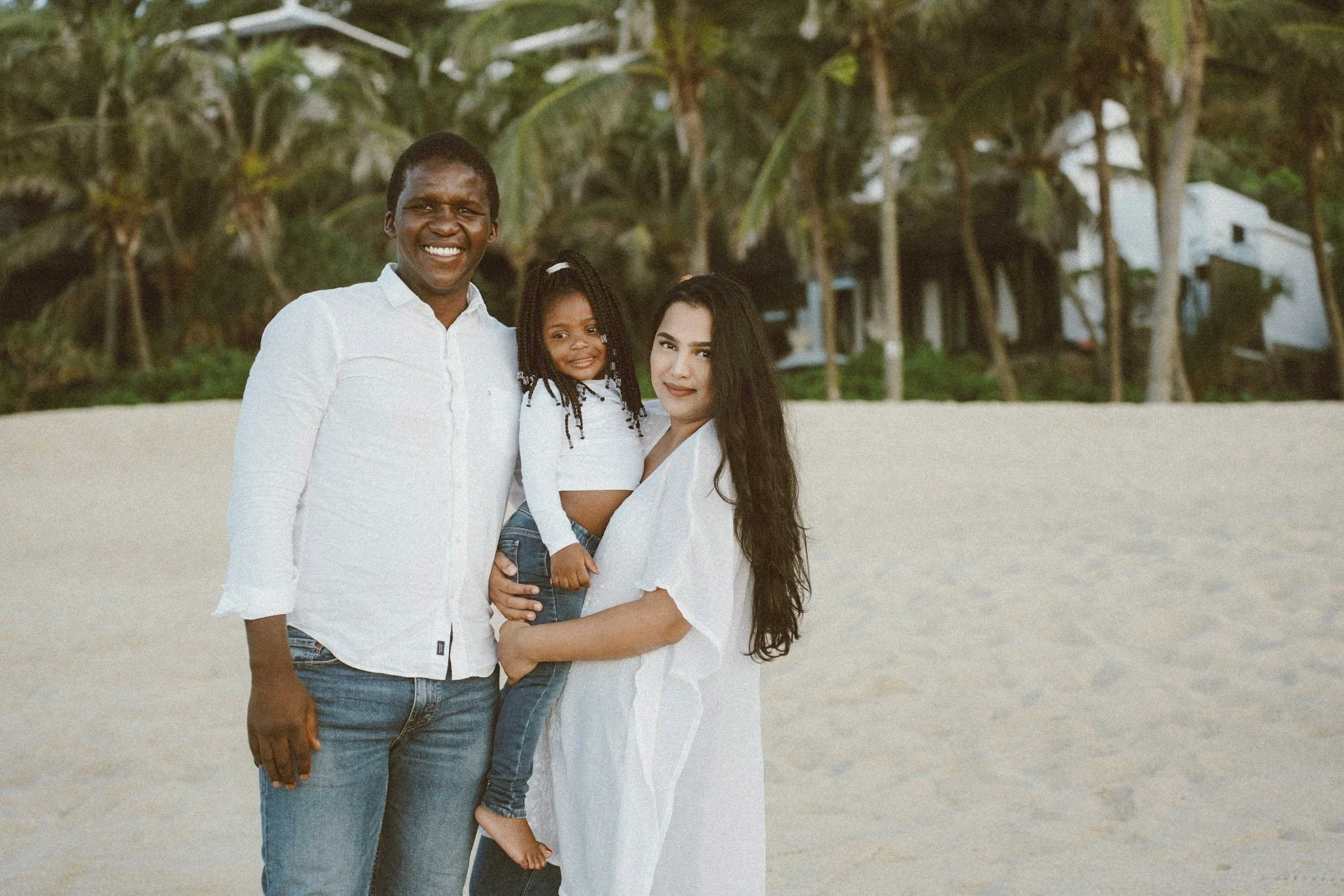 A multicultural family of three, including a man, woman, and young girl, standing on a beach with trees in the background during daytime. They are smiling and dressed in casual white and denim clothing.