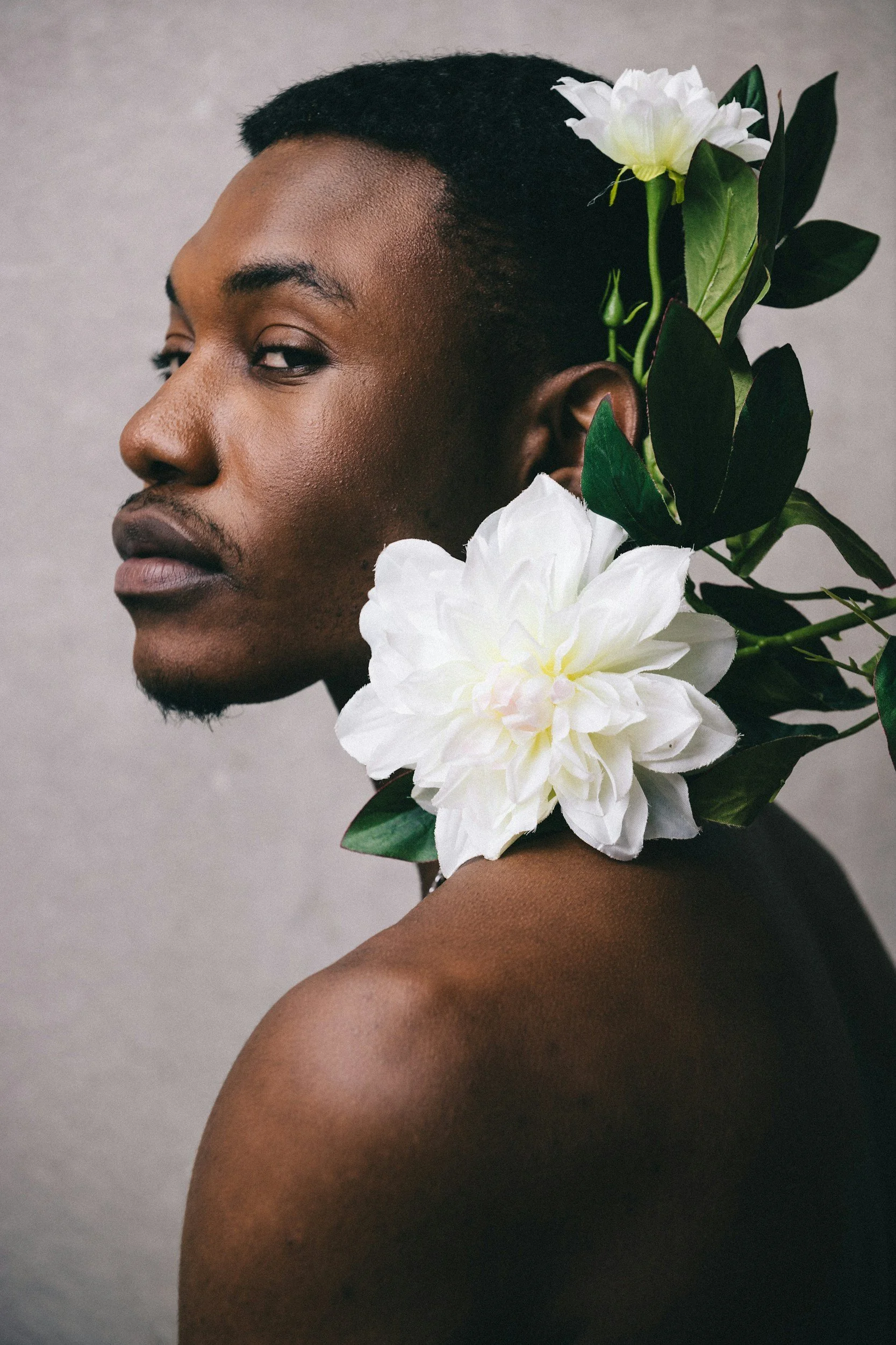 Side profile of a shirtless Black man with flowers behind his ear, against a neutral gray background.