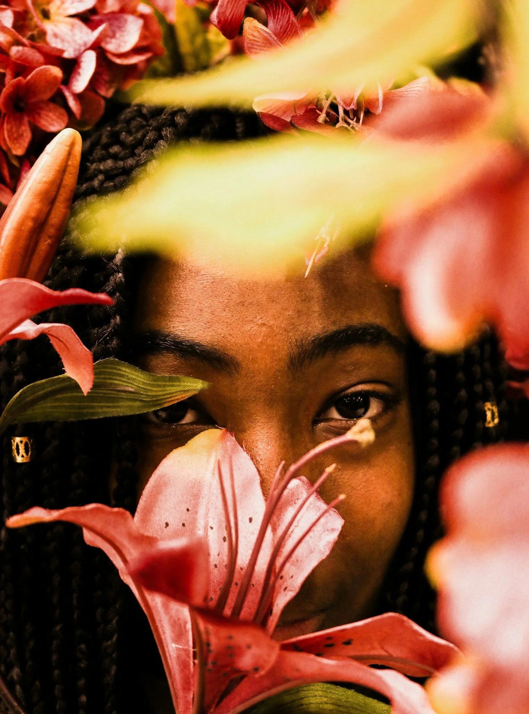 Close-up of a person with dark curly hair, partially obscured by pink lilies and red flowers, with a focus on their eyes and forehead.