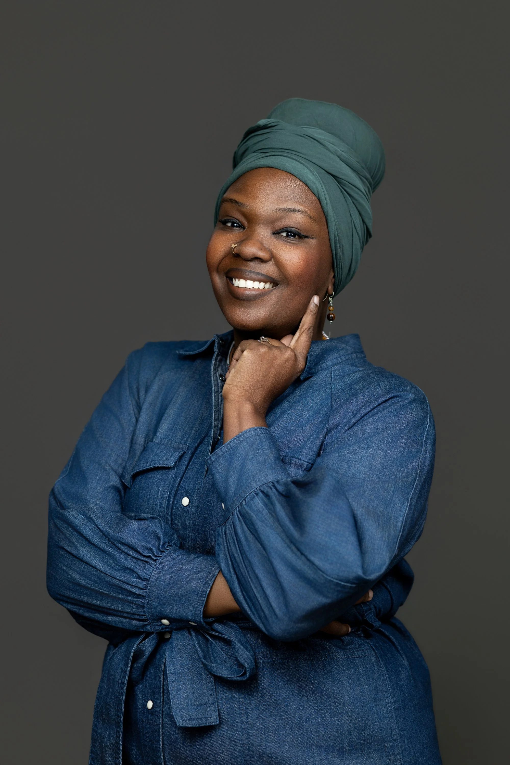 Portrait of a woman with dark skin, wearing a green headwrap and a denim shirt, smiling and touching her cheek, against a gray background.