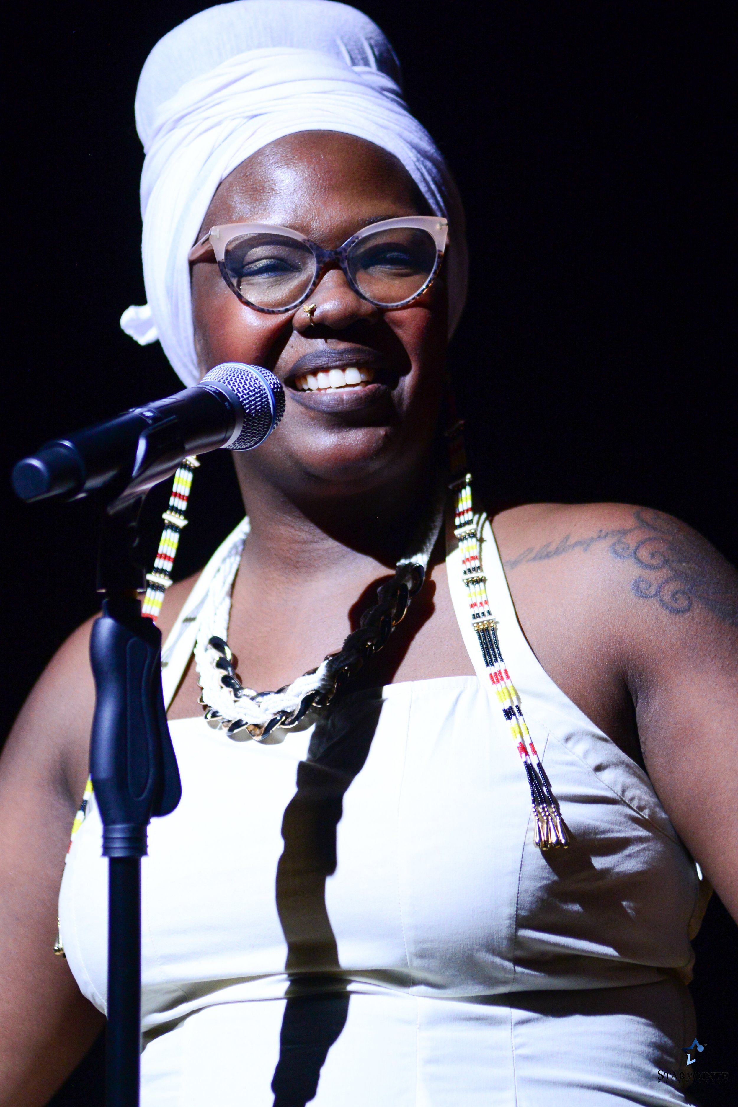 A woman smiling and speaking into a microphone on stage, wearing glasses, a headwrap, and beaded jewelry, with a tattoo visible on her upper arm.