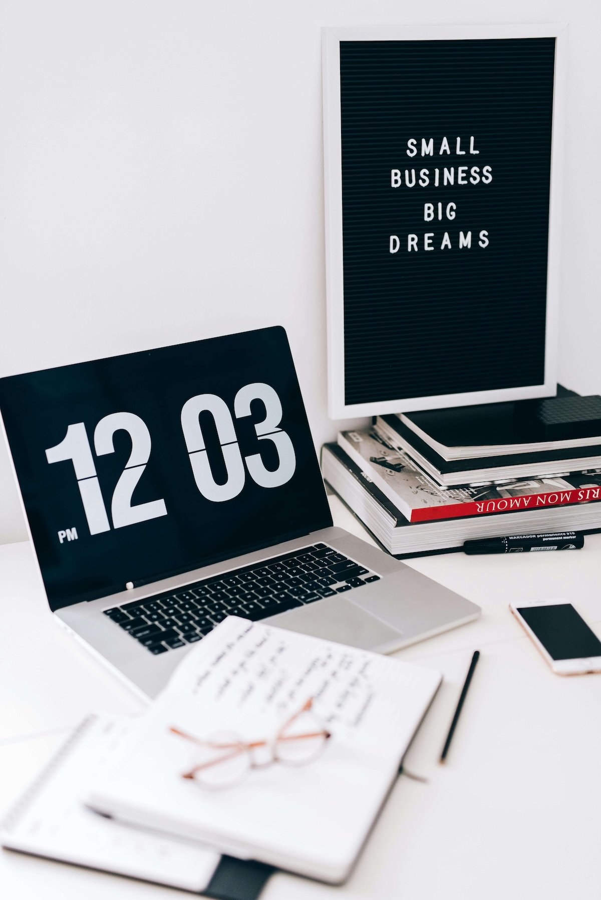 Office workspace with a laptop displaying 12:03 PM, a letter board with 'Small Business Big Dreams,' stacked books, a smartphone, notebook, and glasses.