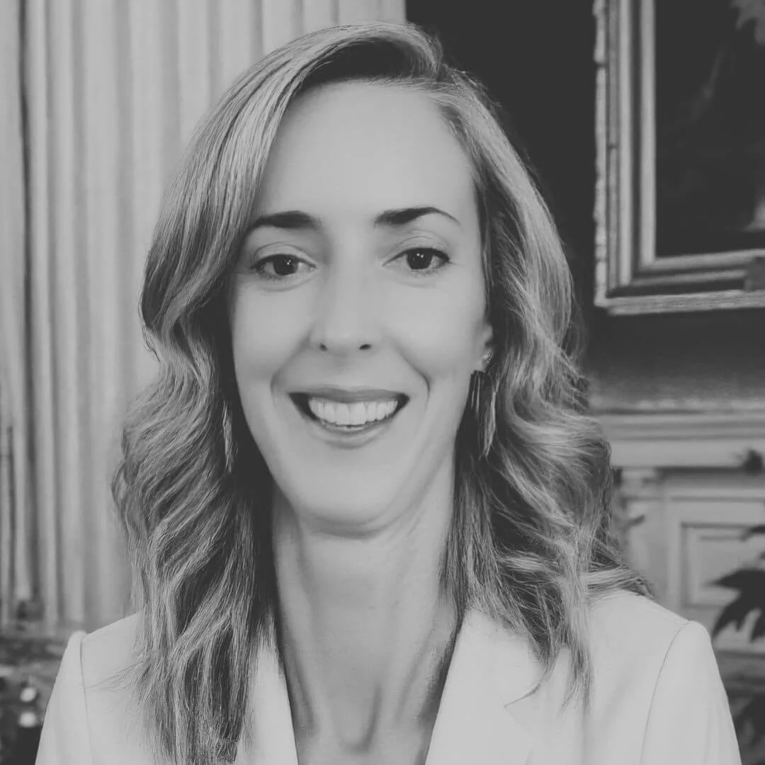 Black and white photo of a smiling woman with wavy hair wearing a white jacket, standing against an ornate interior background.