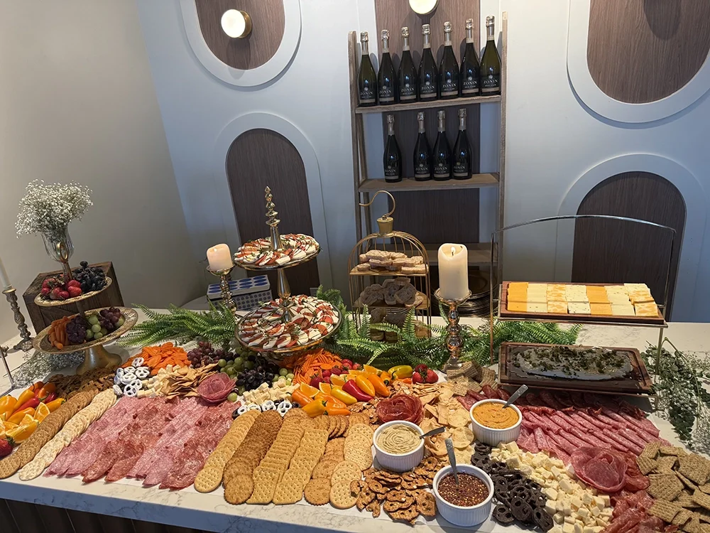An elaborate buffet table with assorted meats, cheeses, crackers, fresh fruits, and desserts, including cookies and chocolates. Beer bottles are on a shelf in the background, and there are decorative candles and greenery on the table.