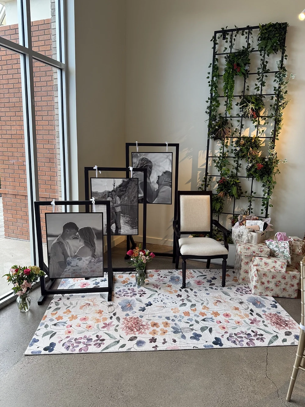Display of black-and-white portrait photographs of a couple, placed on black frames and supported by stands, set on a floral-patterned rug. Two flower vases with pink and white flowers are on each side of the display. Behind is a beige wall with a la