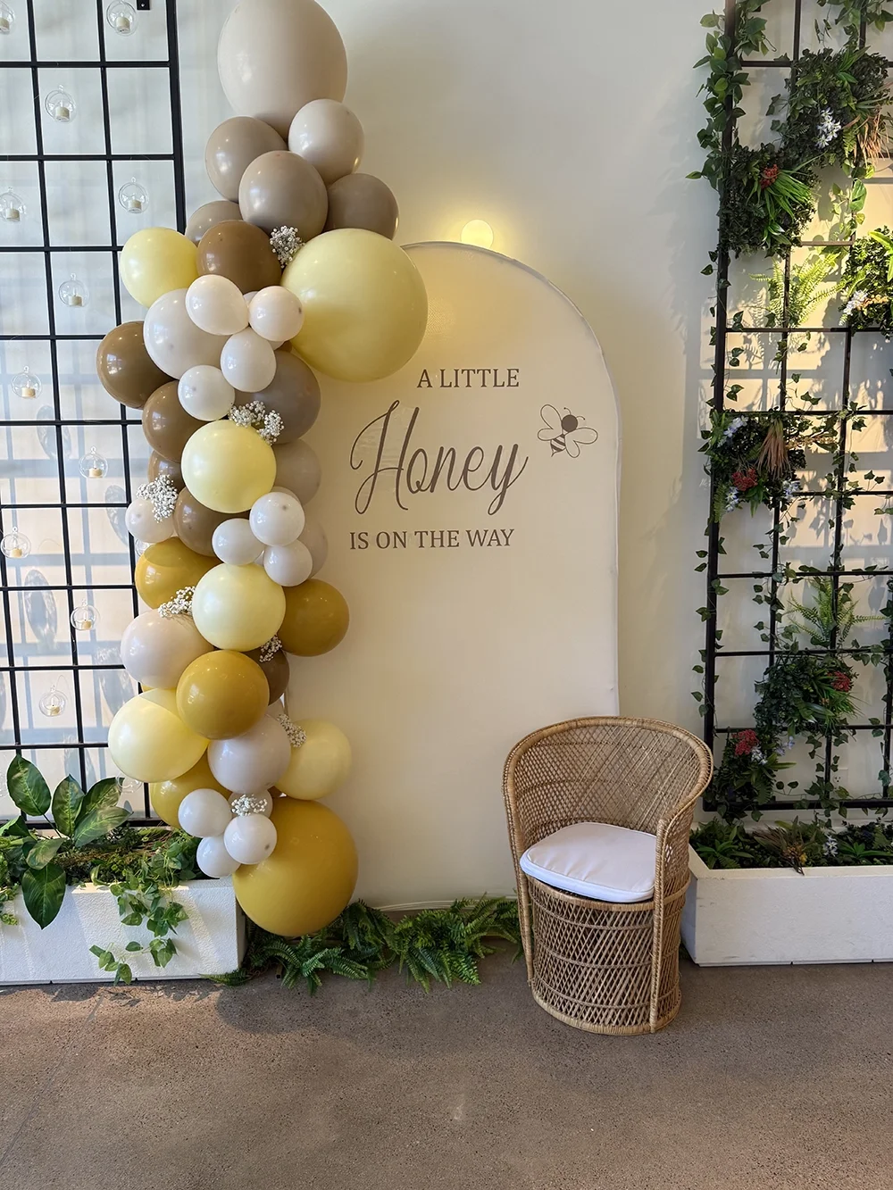 Decorative setup with a balloon arch featuring beige, yellow, and white balloons, a wall sign reading "A Little Honey is on the Way" with a bee illustration, greenery, and a wicker chair with a white cushion.