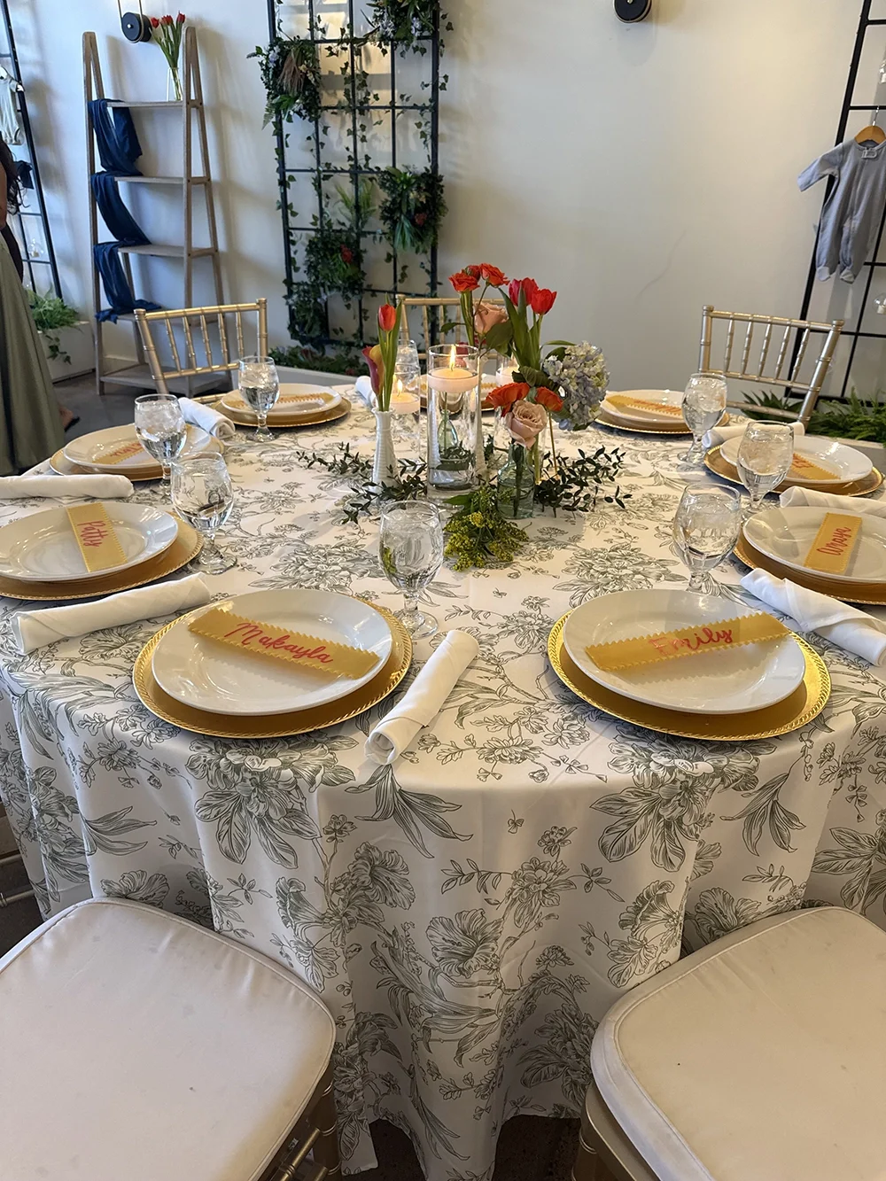 A round dining table set for a formal meal with a floral tablecloth, gold-rimmed plates, white napkins, water glasses, and place cards with handwritten names. In the center, there is a floral arrangement with pink, red, and white flowers and candles.