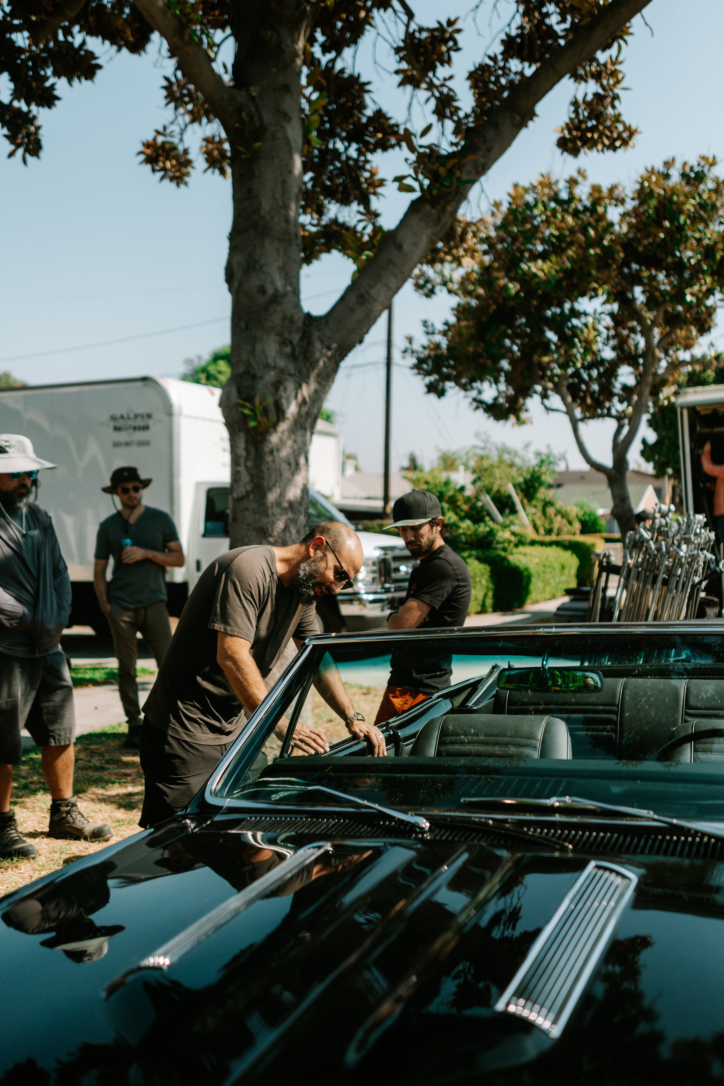 Group of people standing around a black convertible car with trees and white trucks in the background. Powerful still from Courage by Eric Hernandez—a modern story of resilience, healing, and tradition