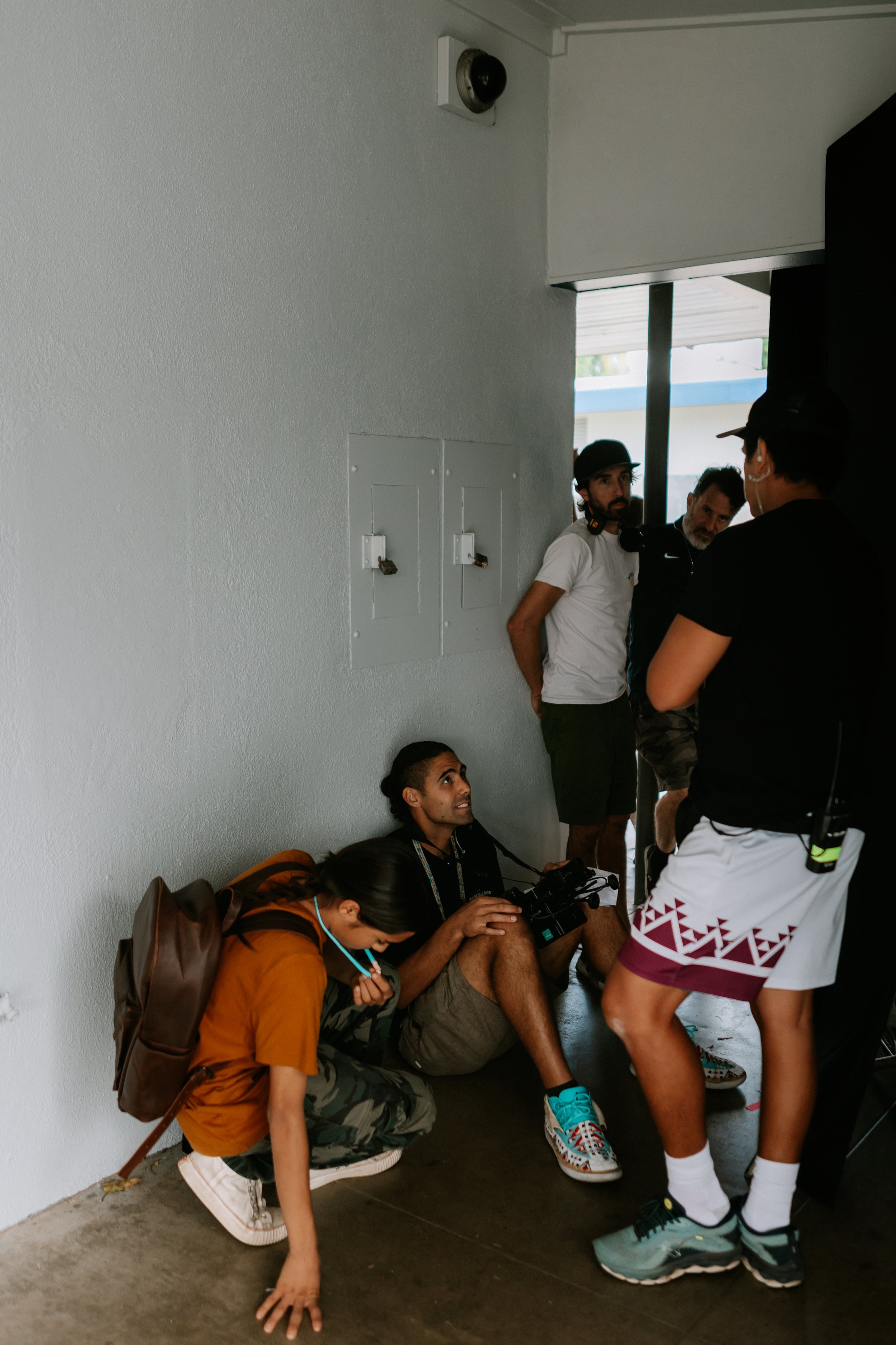 A group of people in a hallway, with some sitting and others standing. One person is sitting with a camera, and another is wearing a backpack. Powerful still from Courage by Eric Hernandez—a modern story of resilience, healing, and tradition
