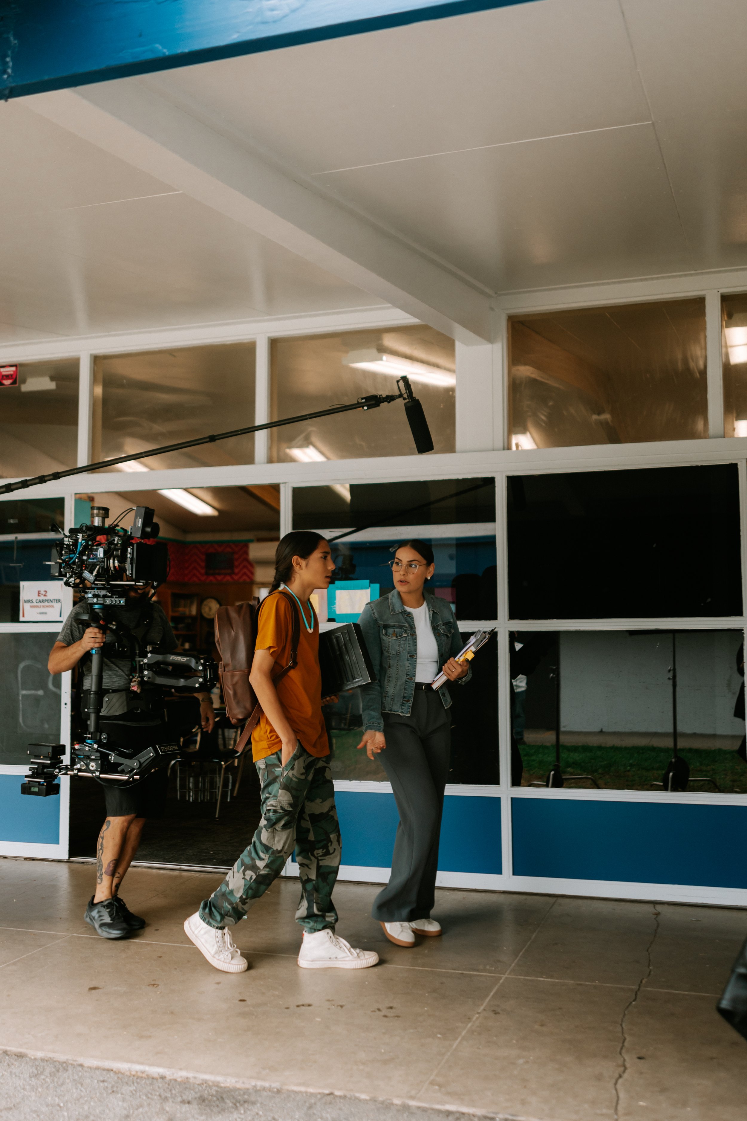 Film set with a camera operator using a stabilizer rig to film two people walking outside a building, one holding a folder and the other wearing a backpack. A boom microphone is visible overhead.