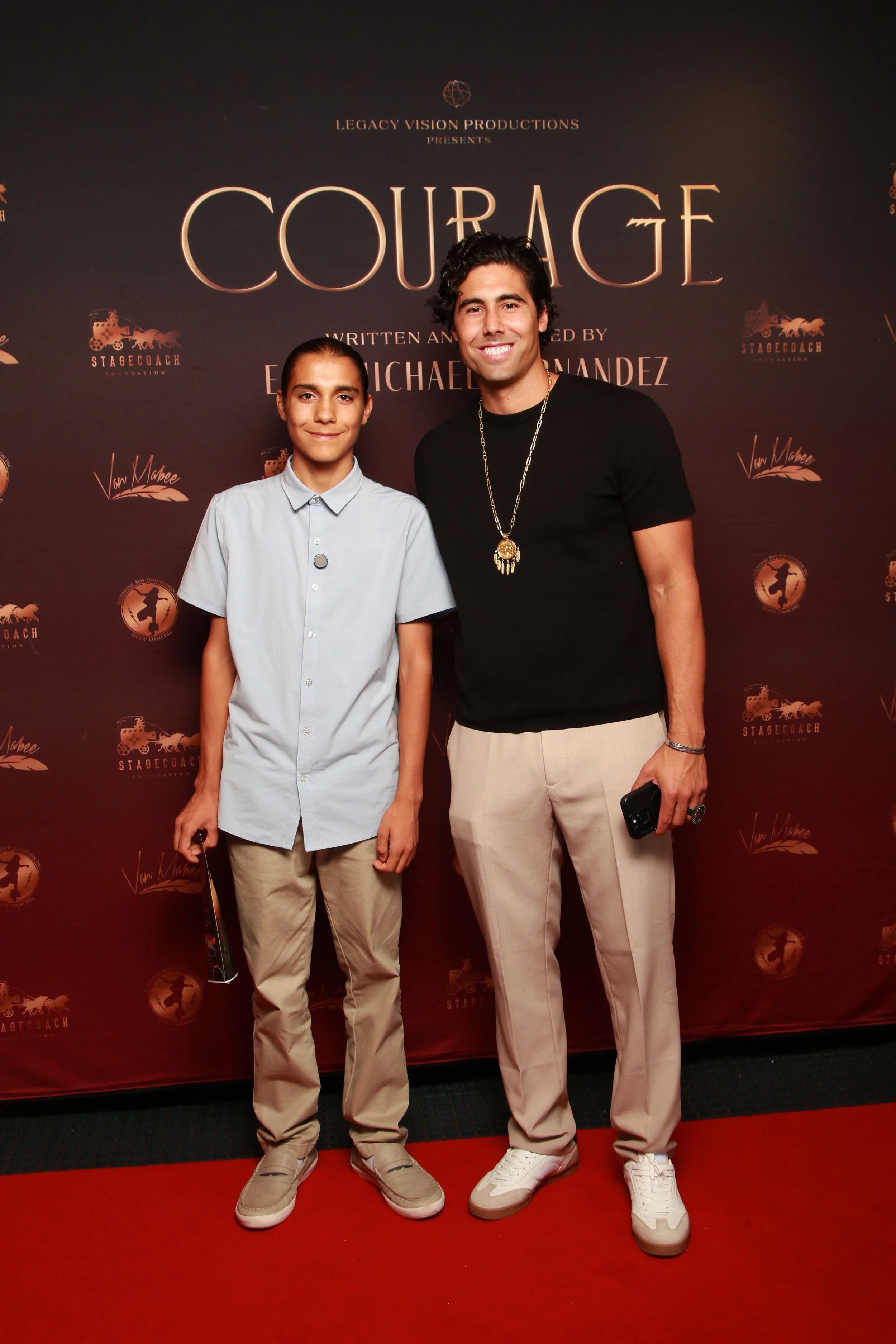 Two men standing on a red carpet in front of a backdrop for the play 'Courage'. One is a young girl wearing a light blue button-up shirt and beige pants, holding a booklet. Courage Screening premier, Eric Hernandez