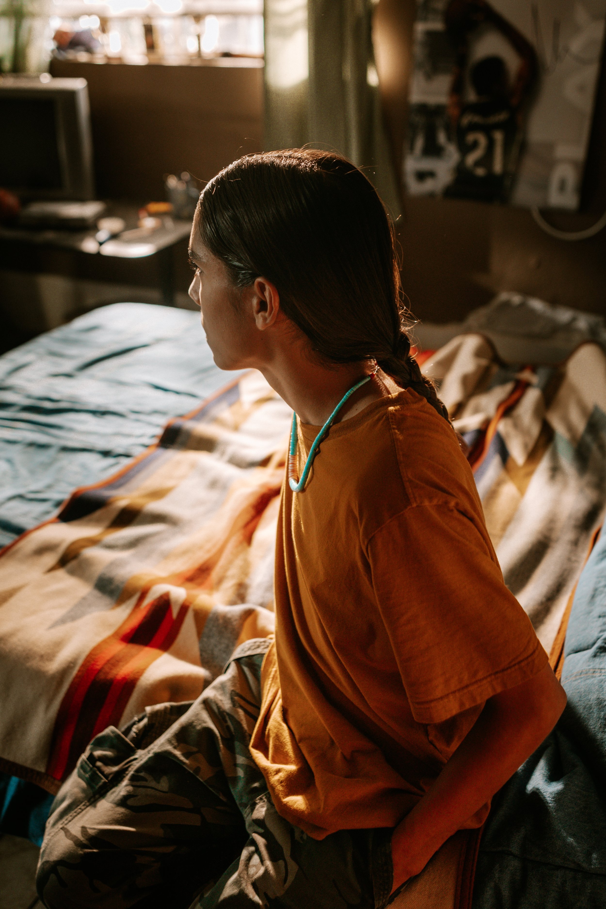 A person with long hair sits on a bed in a dimly lit room. They are wearing an orange t-shirt and camouflage pants. The room has a blanket with geometric patterns, a poster on the wall, and a desk with small items.