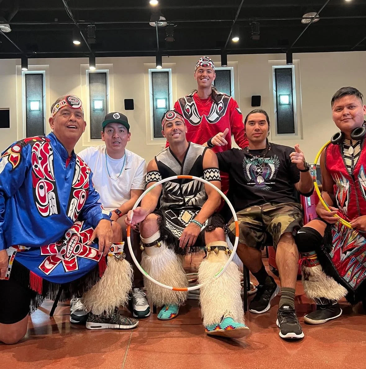 Six men dressed in traditional Indigenous attire and modern casual clothing, posing together in an indoor setting with a high ceiling and wall-mounted lights.