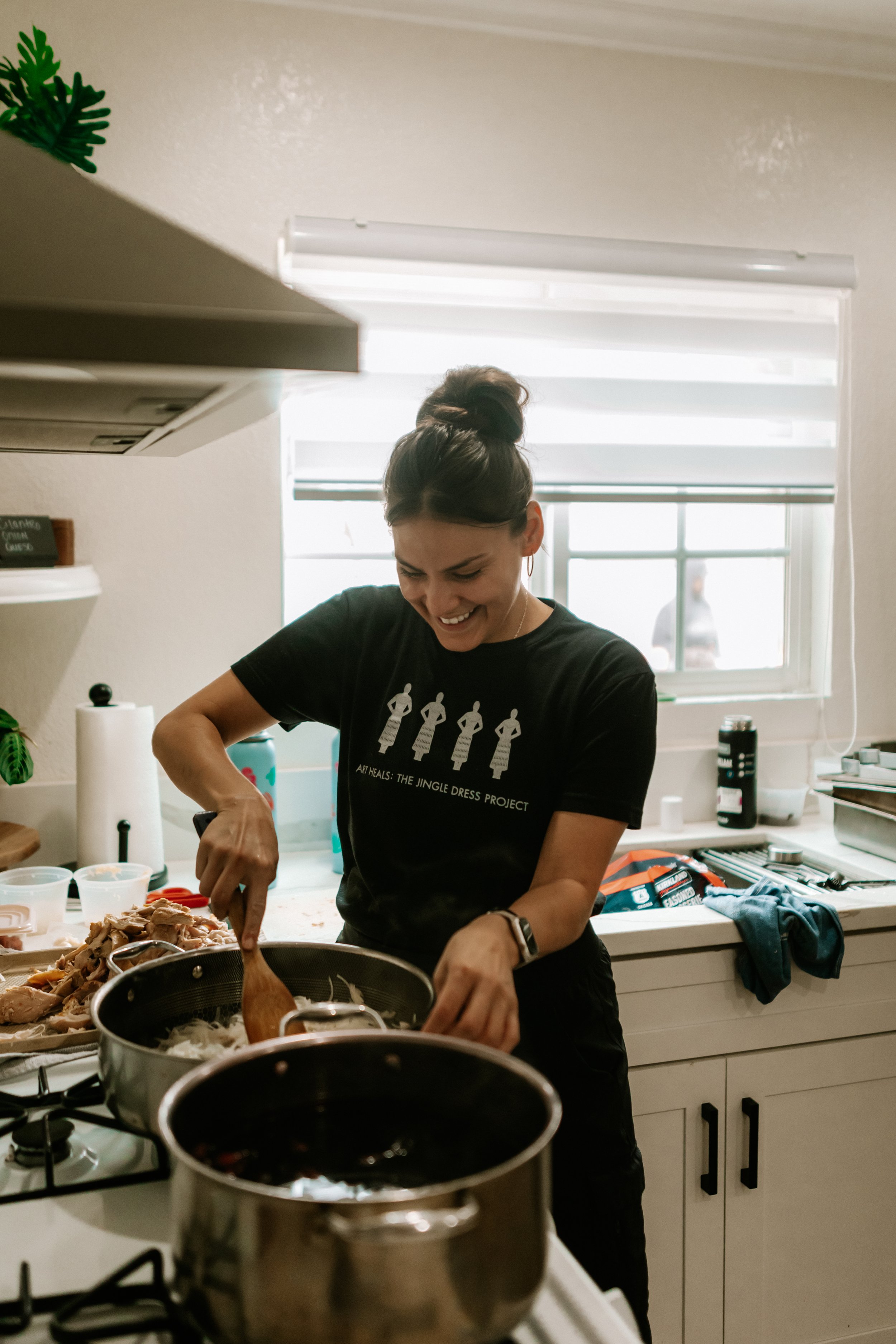A person smiling while cooking in a kitchen, stirring a pot on the stove with a wooden spoon, wearing a black t-shirt with "Art Heals: The Jingle Dress Project" printed on it. There are ingredients and kitchen items on the counter. Chef Pyet DeSpain.