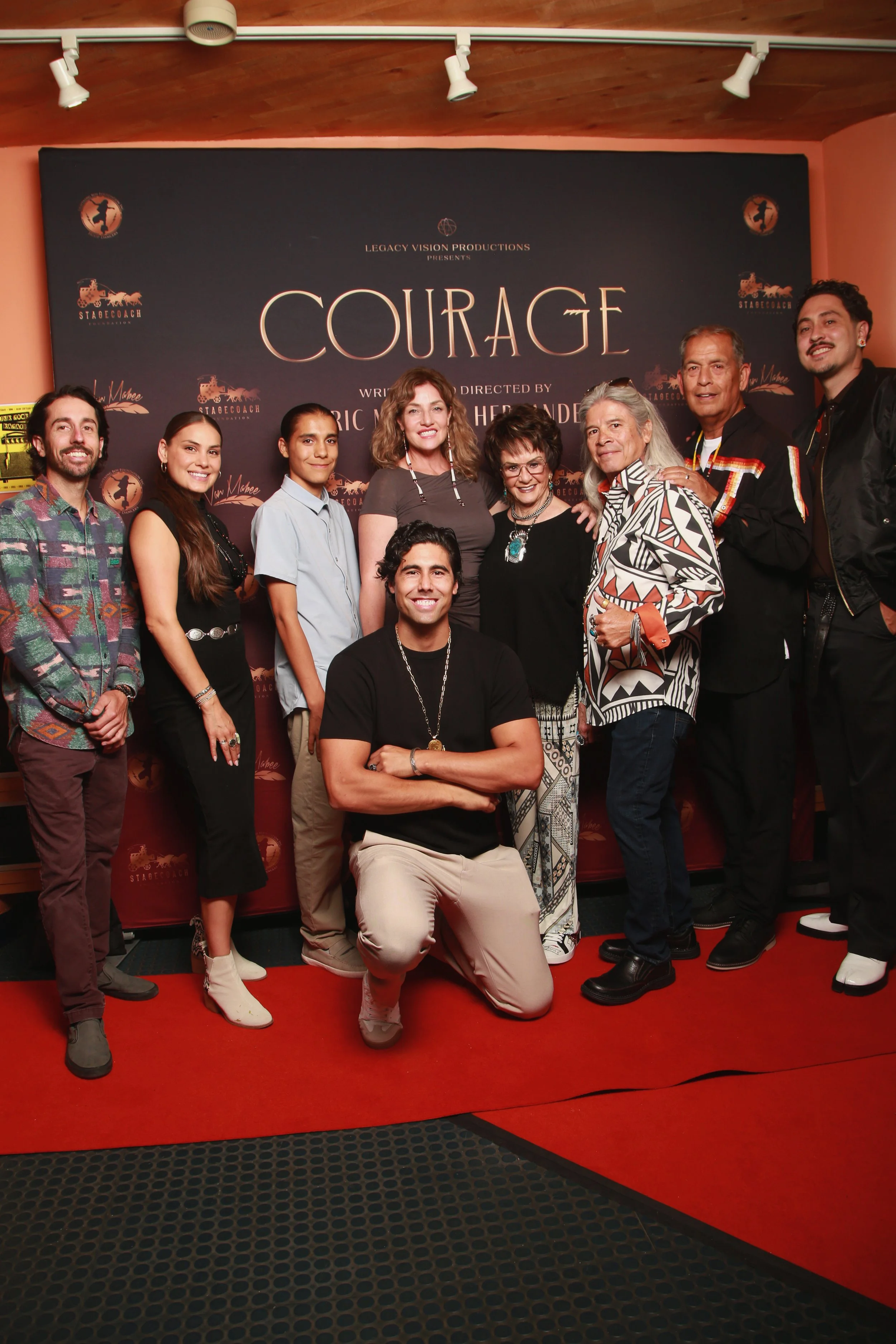 Group of nine people posing in front of a movie poster for 'Courage' at a film event. Courage Screening premier, Eric Hernandez