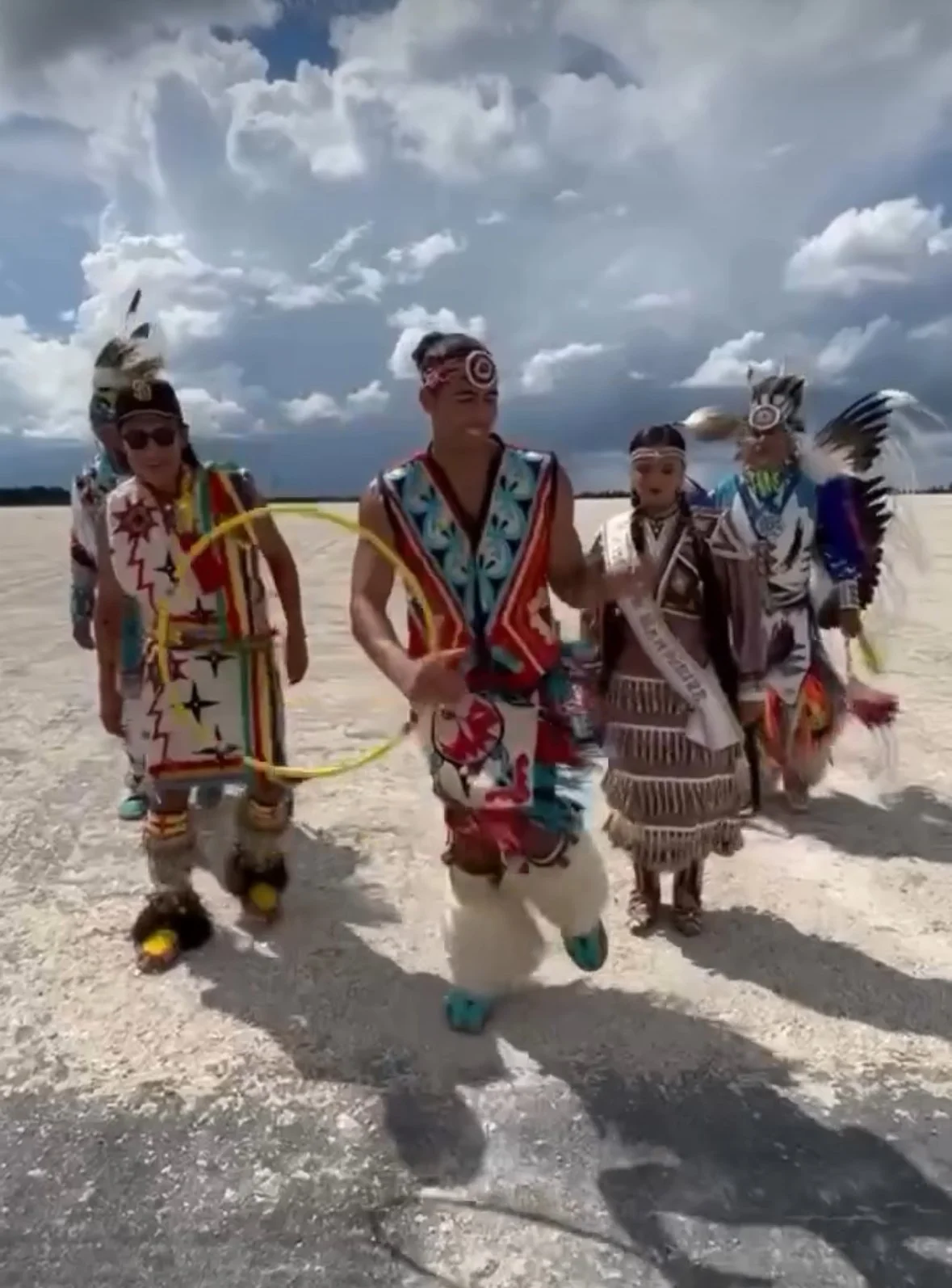 Group of Native Americans in traditional clothing, dancing outdoors on a bright day with clouds in the sky.