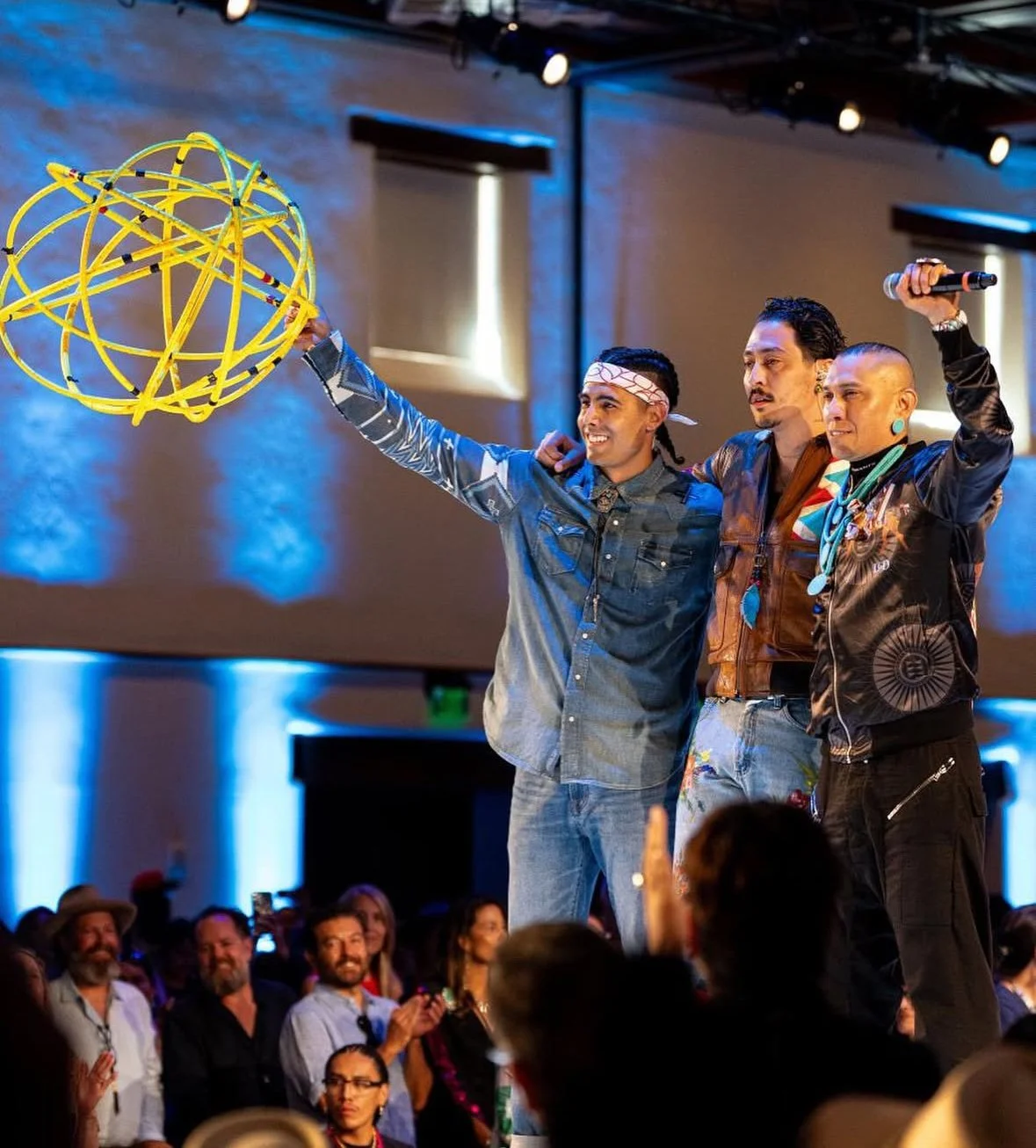 Three men on stage, one holding a yellow spherical sculpture, and another holding a microphone, with an audience in the background.