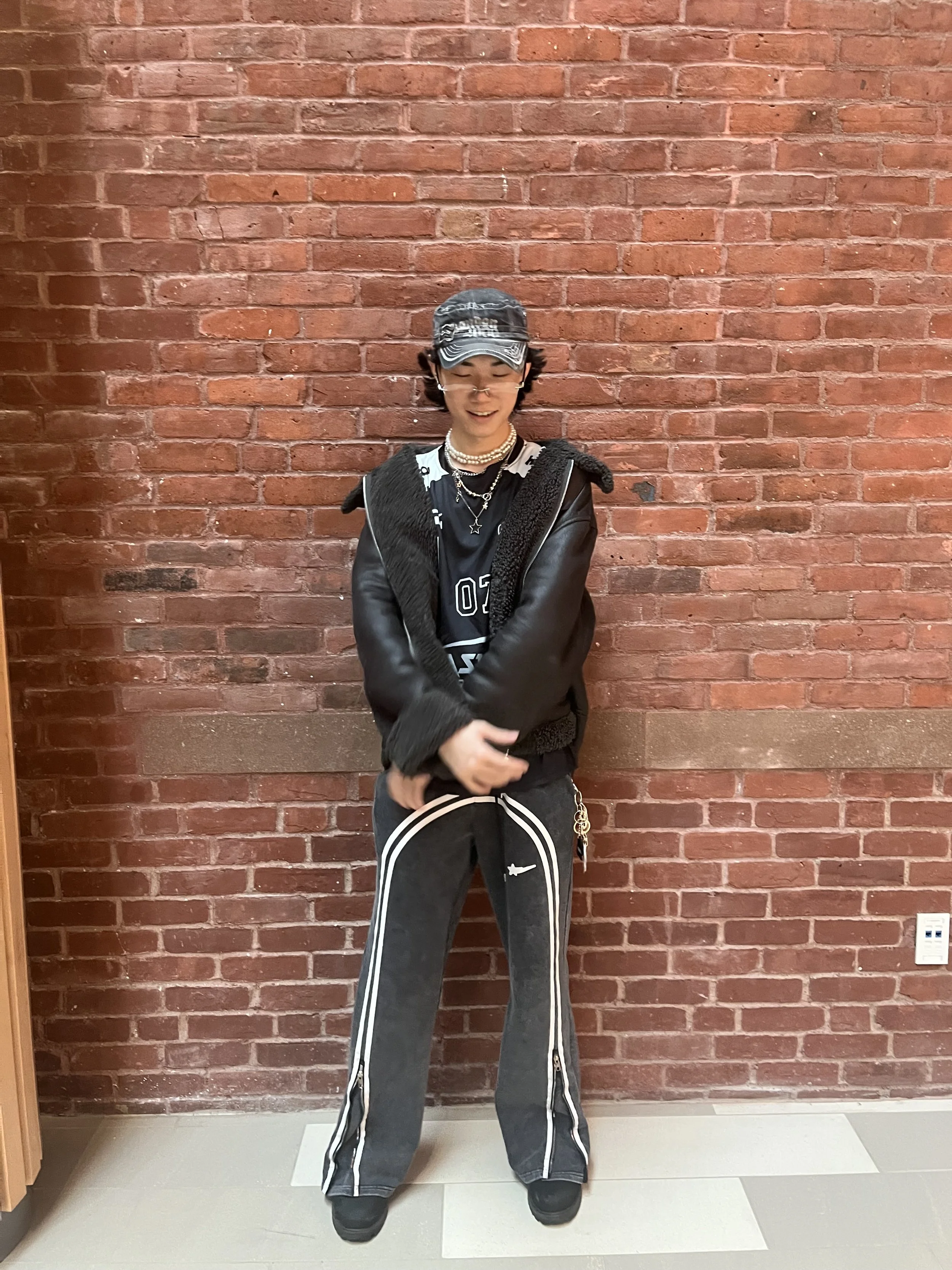 Young woman wearing a black cap, layered necklaces, pearl collar, black graphic shirt, black jacket with fur lining, black pants with white stripe details, and black shoes, standing in front of a red brick wall.