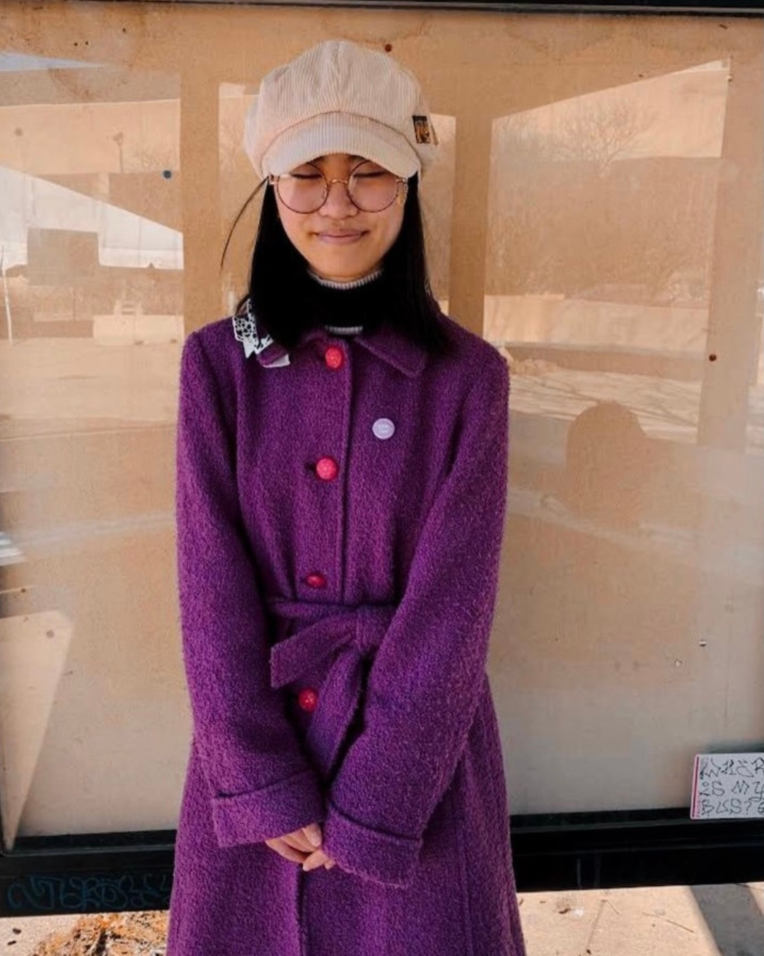 A young woman wearing glasses, a beige cap, and a purple coat with colorful buttons, standing outdoors in front of a glass window with reflections of trees and buildings.