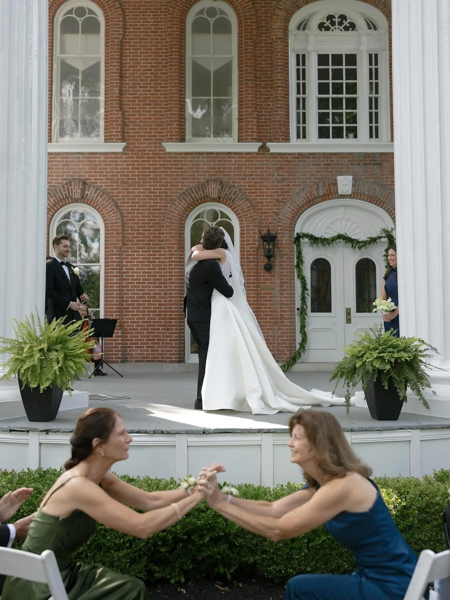 A wedding ceremony with a groom and bride embracing on an outdoor platform, with bridesmaids and groomsmen in the background, and guests sitting and dancing in front of the stage at a sunny outdoor venue with a brick building in the background.