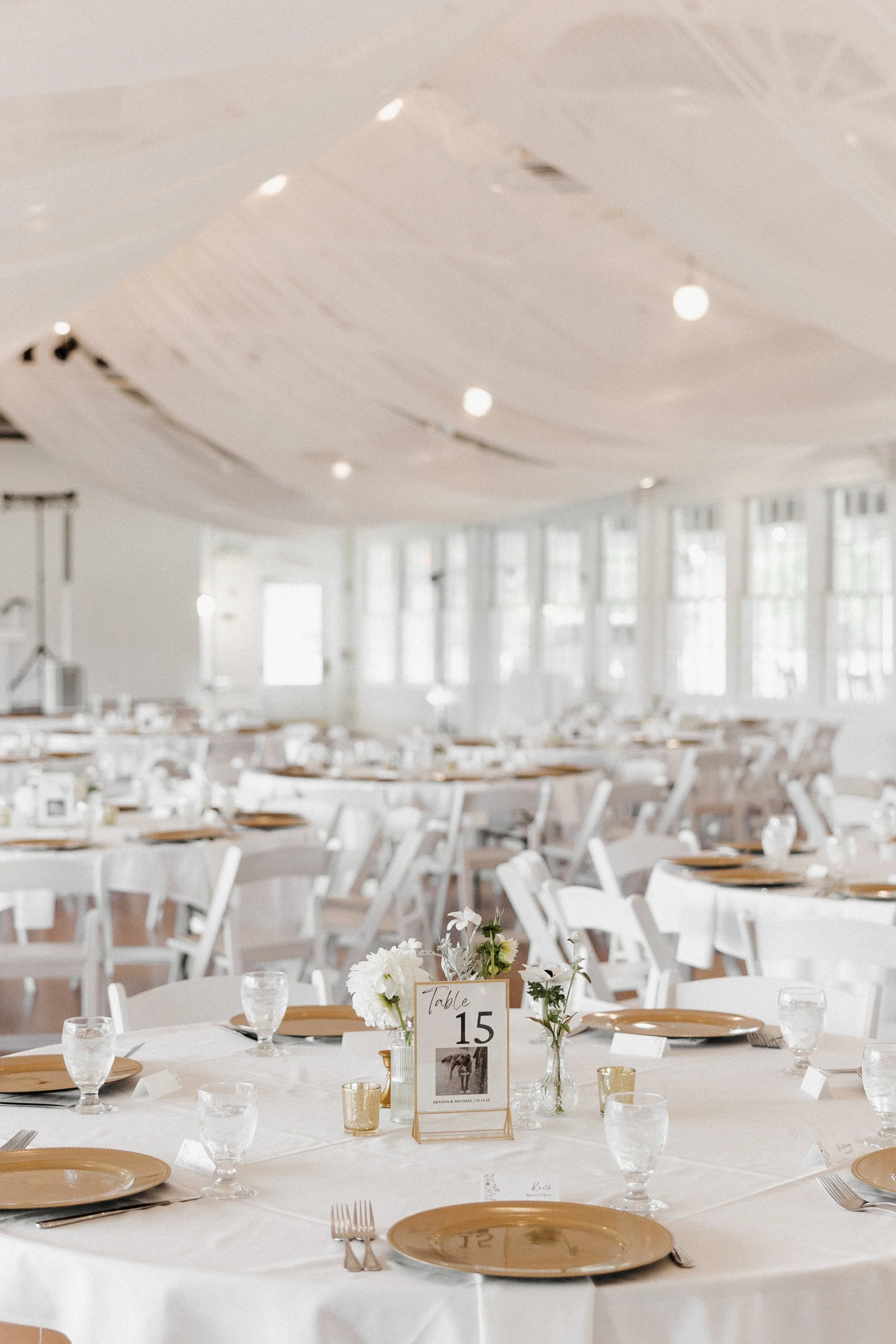 A large banquet hall decorated for a wedding reception with round tables covered in white tablecloths, each set with gold chairs, plates, silverware, and glasses. Centerpieces feature white flowers and a table number card for table 15. The room is br
