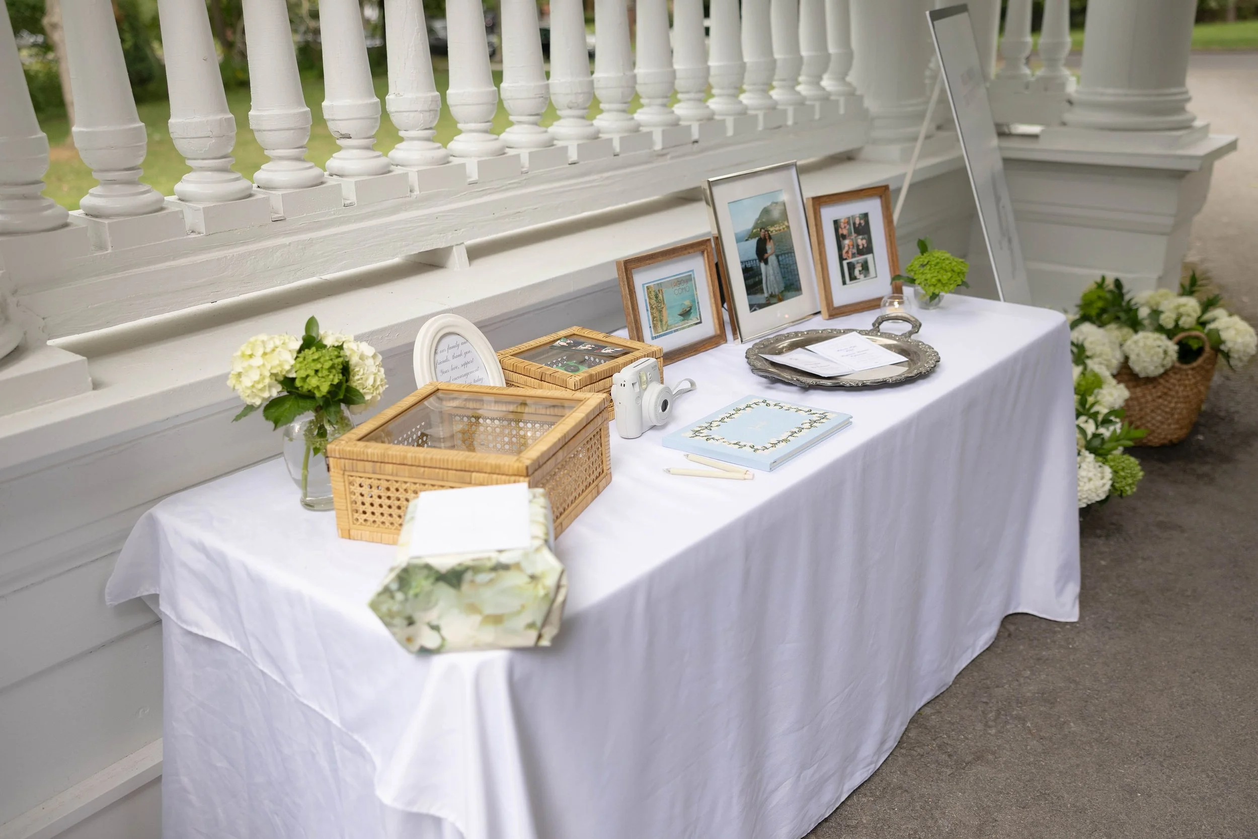 Table with framed photographs, flowers, a camera, and decorative items, set against a white porch railing and greenery background.
