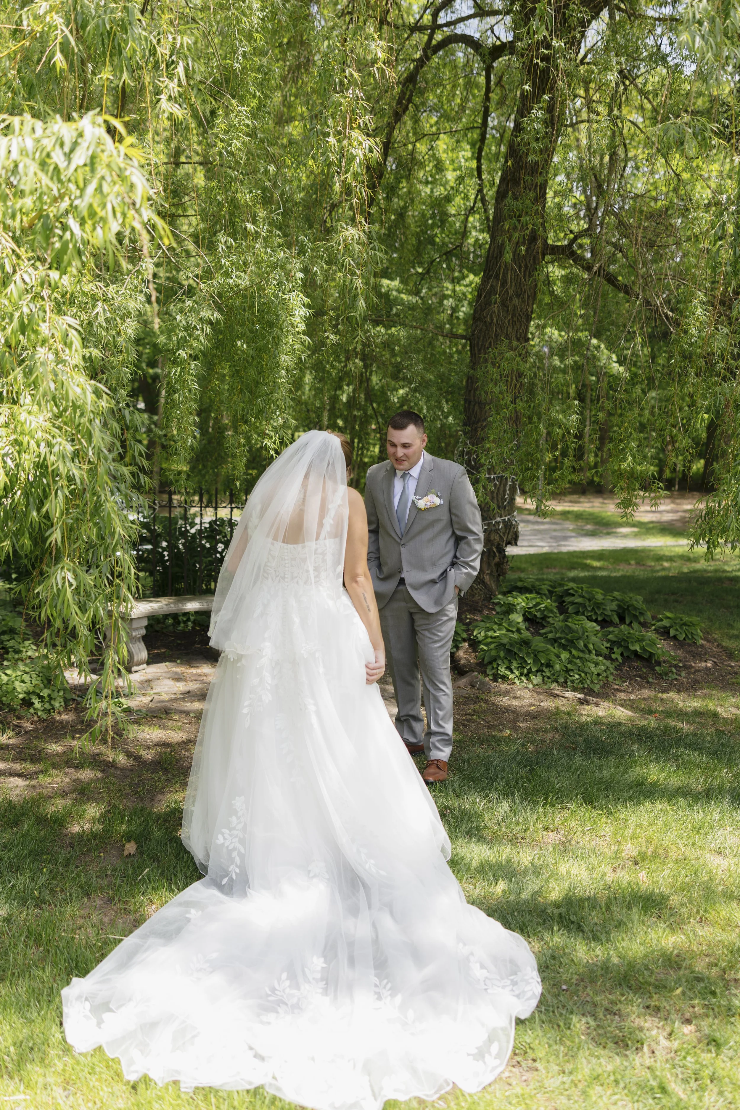 A bride and groom standing outdoors under a tree, facing each other on a sunny day. The bride is in a white wedding gown with a long veil, and the groom is in a gray suit with a boutonniere.