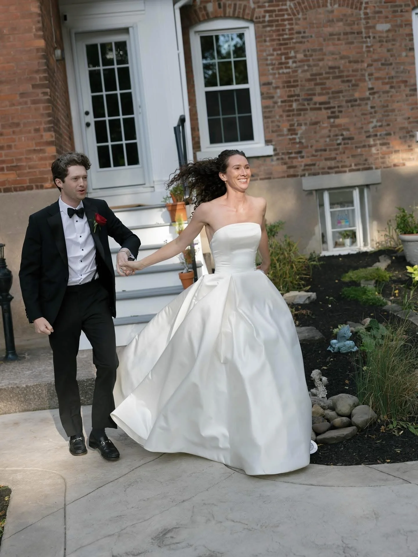 A bride in a strapless white wedding gown smiling while walking outside, holding a groom's hand, in front of a brick house with garden plants.