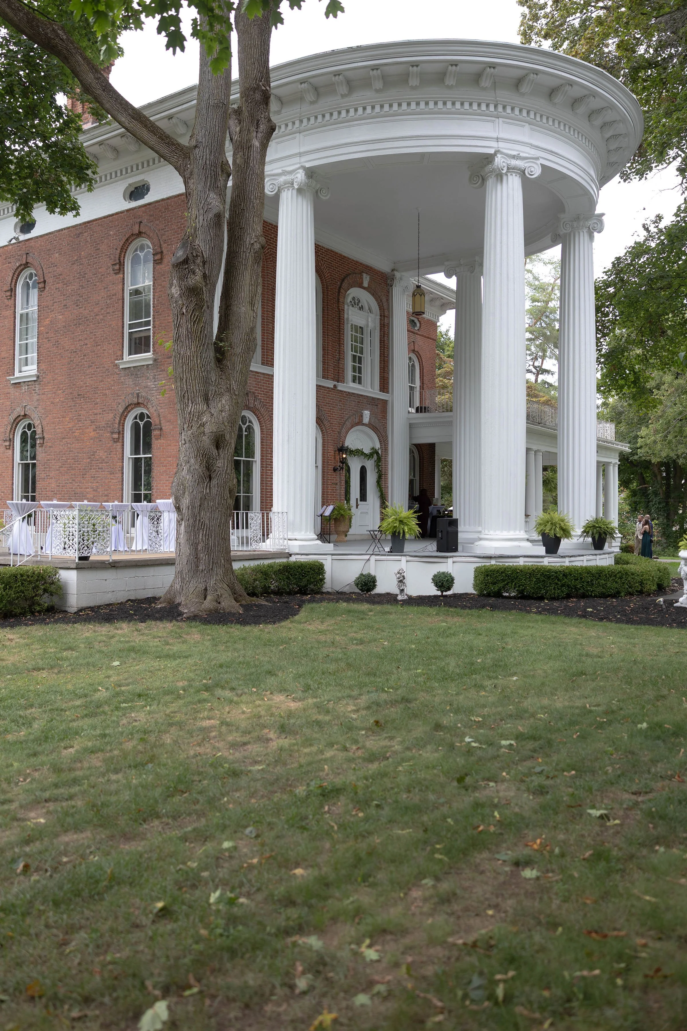 A large, historic brick house with a white portico and four tall, white columns. There are trees and greenery in the yard, with a table set up outside and a person visible near the entrance.