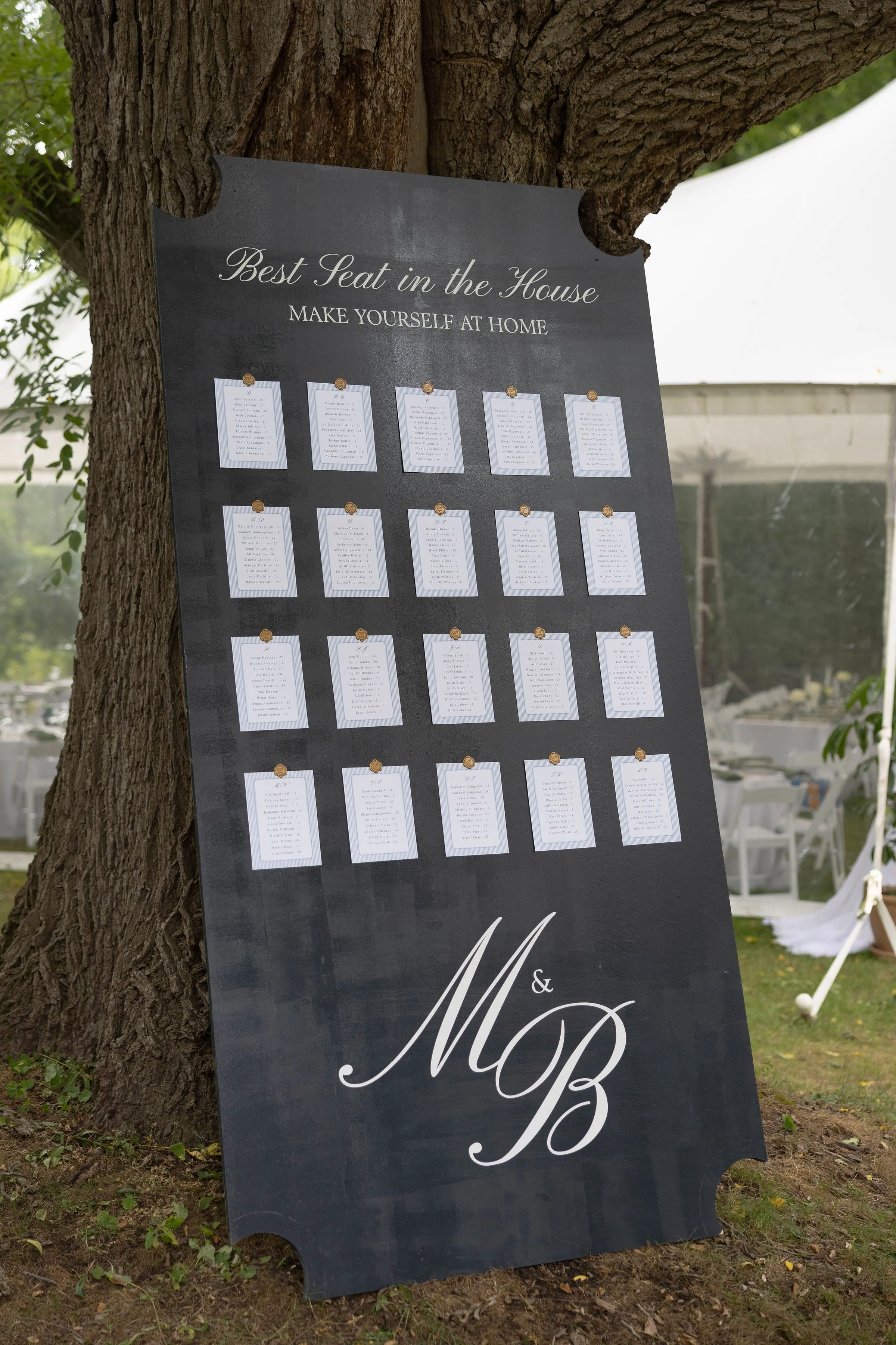 A large black wooden sign attached to a tree with small white papers for seating arrangements for a wedding, with the heading 'Best Seat in the House' and 'Make Yourself at Home' written on it.